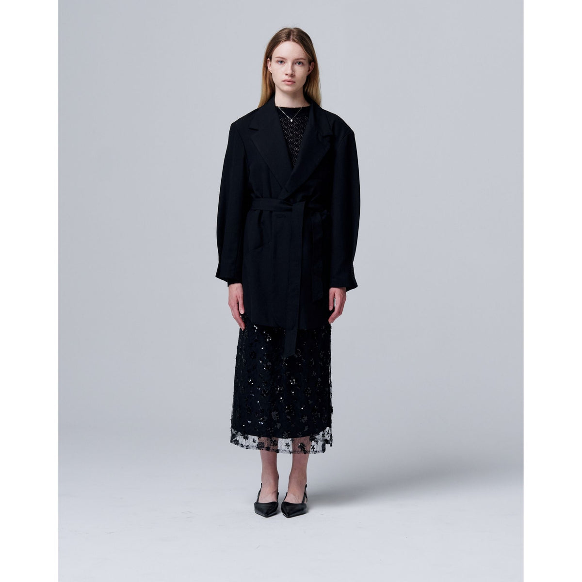 A woman faces forward against a light gray background, wearing ODDPRESENT’s Sequin Beaded Skirt with Floral Detailing under a long black belted coat and black heels.