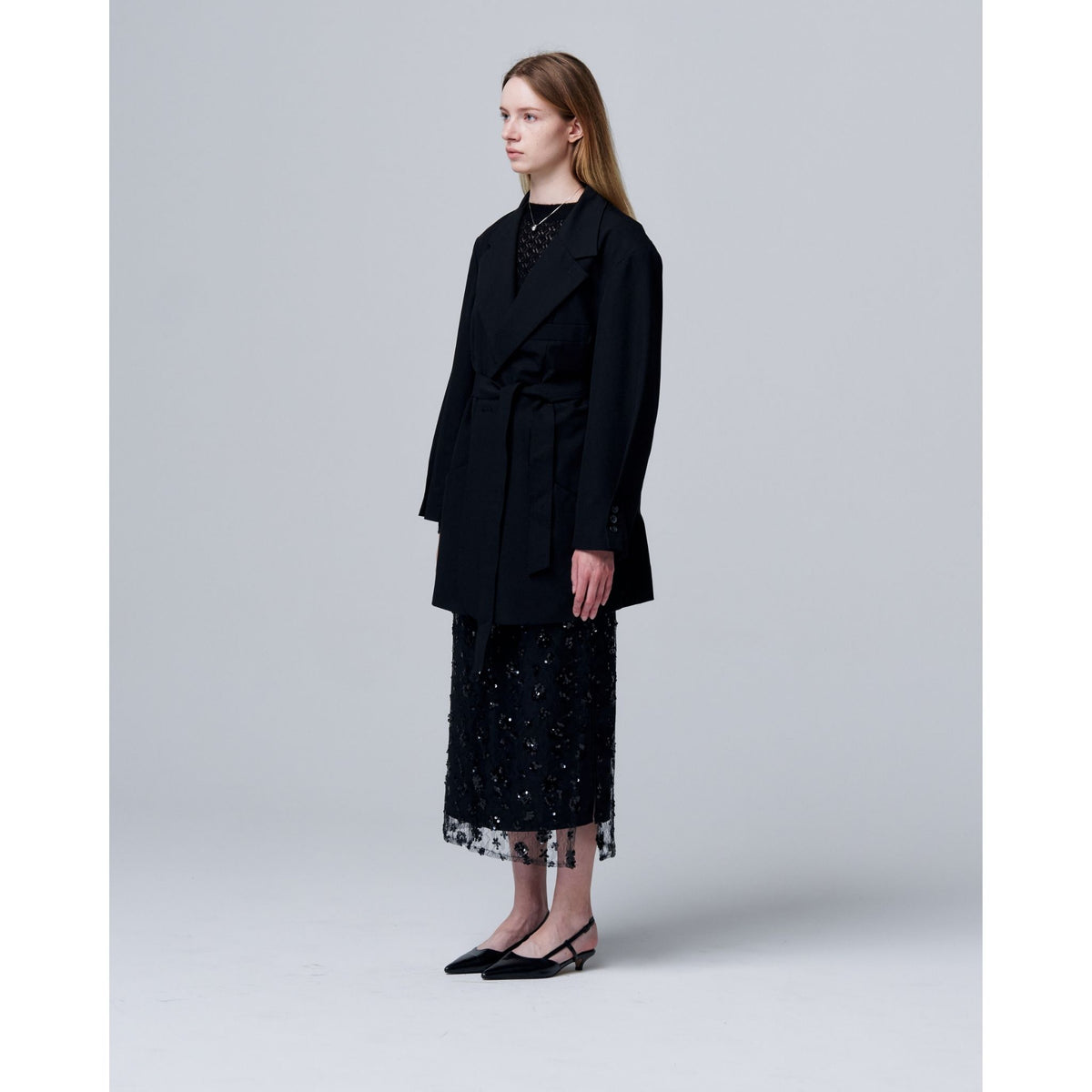 A woman stands against a light gray backdrop wearing ODDPRESENT&#39;s Sequin Beaded Skirt with Floral Detailing, a long black blazer, and black pointed-toe slingback flats—an effortlessly polished evening look. She faces right, expression neutral.