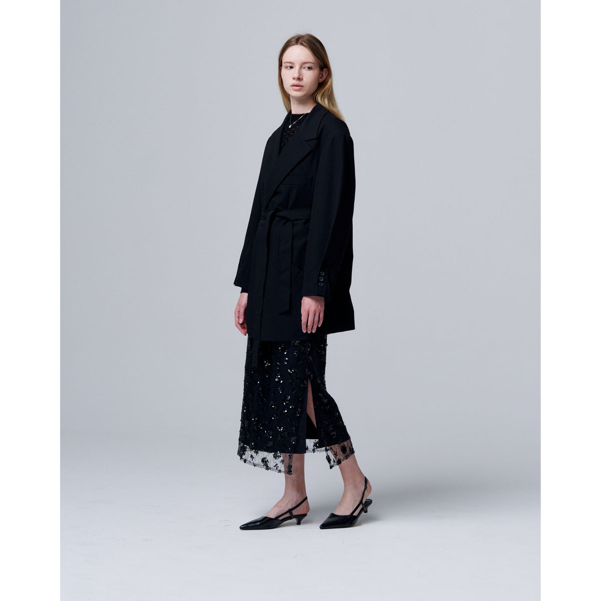 A woman stands in a studio against a light gray background, wearing an ODDPRESENT black belted coat over a sheer, ankle-length dress featuring the Sequin Beaded Skirt with Floral Detailing and black low-heeled slingback shoes.