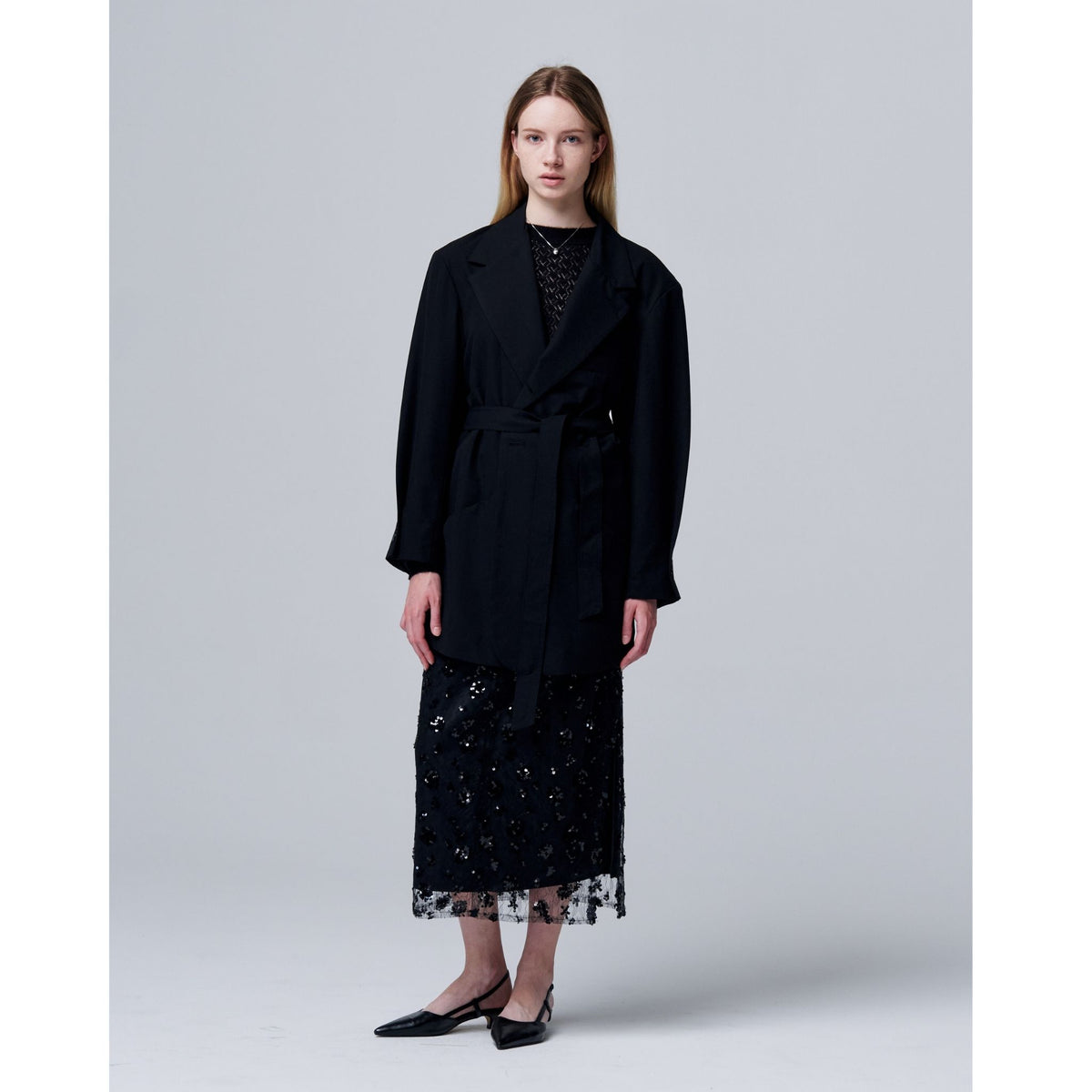 A woman stands against a light gray backdrop, wearing ODDPRESENT’s Sequin Beaded Skirt with Floral Detailing and a black belted coat—perfect for evening events. She completes the look with black pointed-toe flats and straight, shoulder-length hair.