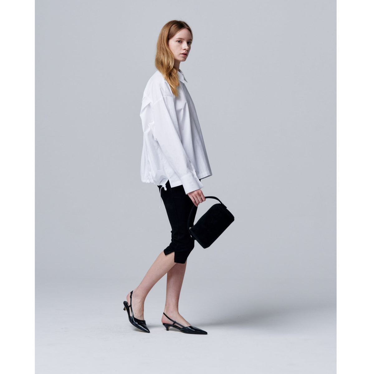 Woman in a white shirt and black pants holding a black handbag on a light gray background
