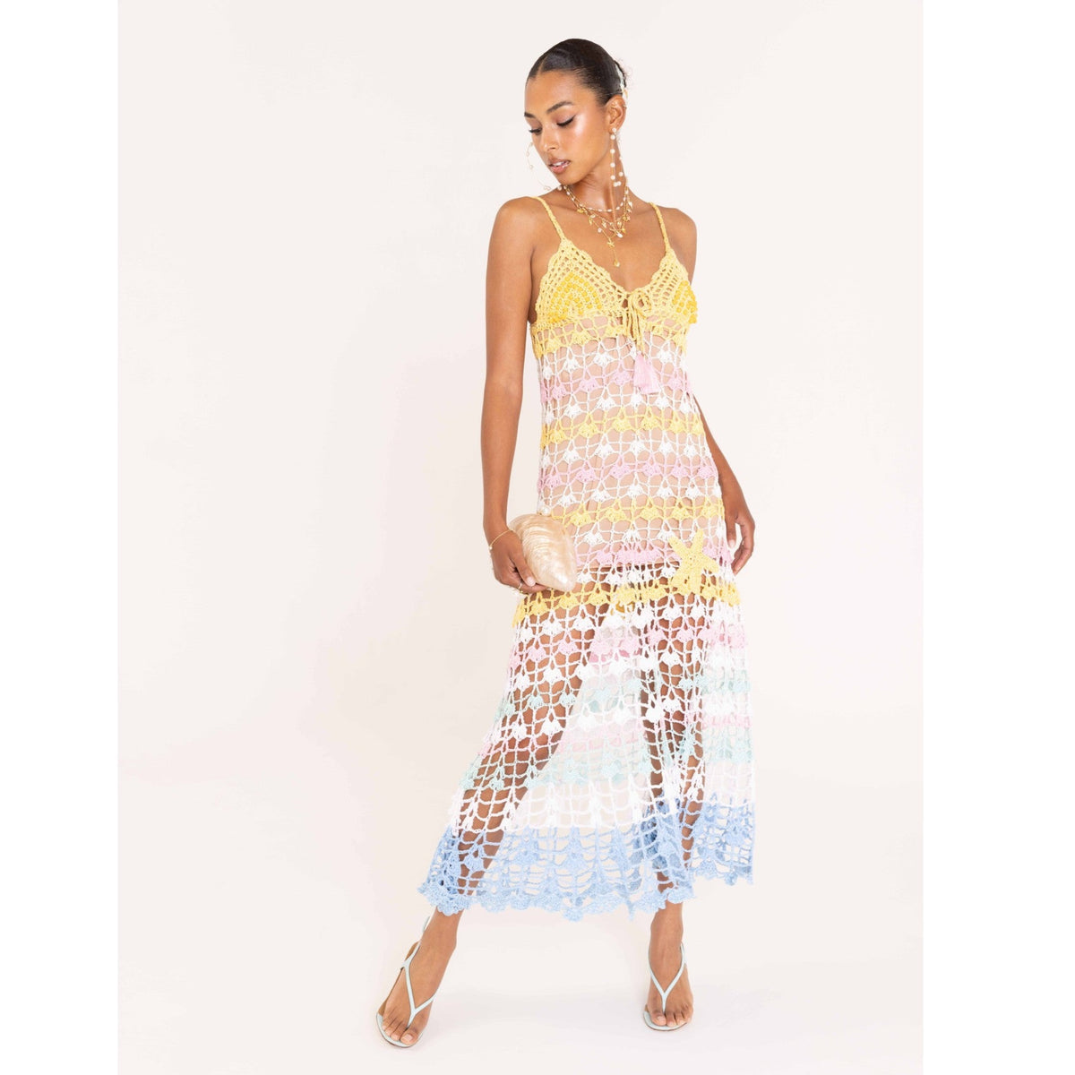 A woman models the ANDREEVA Flower Marine Handmade Maxi Crochet Dress, a colorful sleeveless crochet piece with pastel stripes, paired with strappy heels and a small woven clutch against a white background.