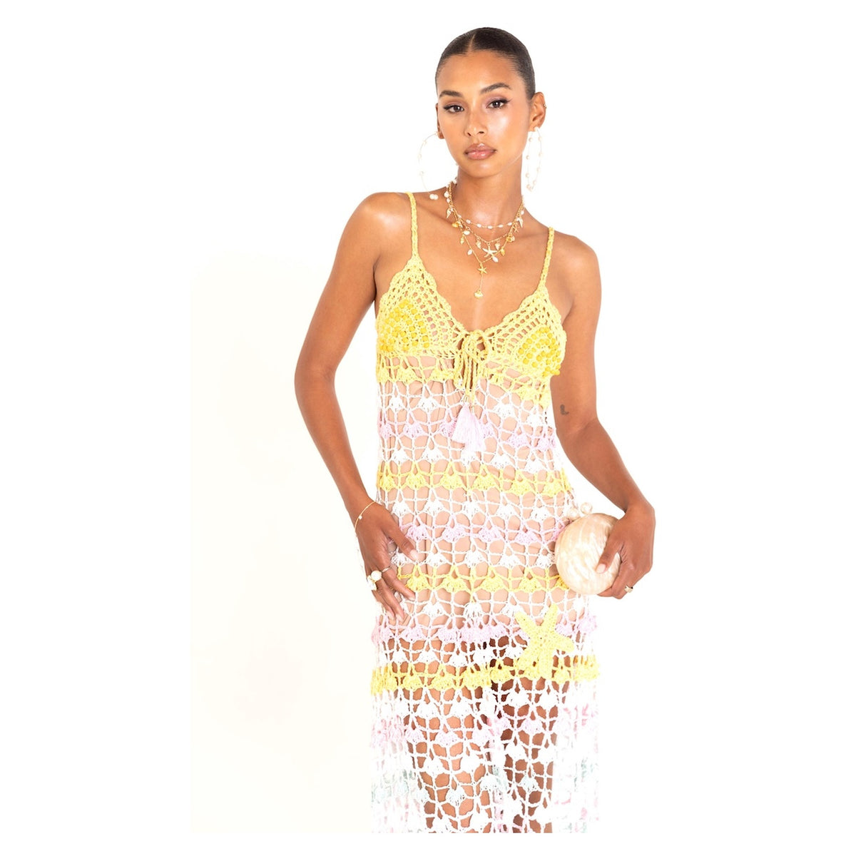 A woman models the ANDREEVA Flower Marine Handmade Maxi Crochet Dress, a sleeveless, multicolored piece with yellow accents and floral patterns. She accessorizes with a round clutch, hoop earrings, layered necklaces, and rings against a white background.