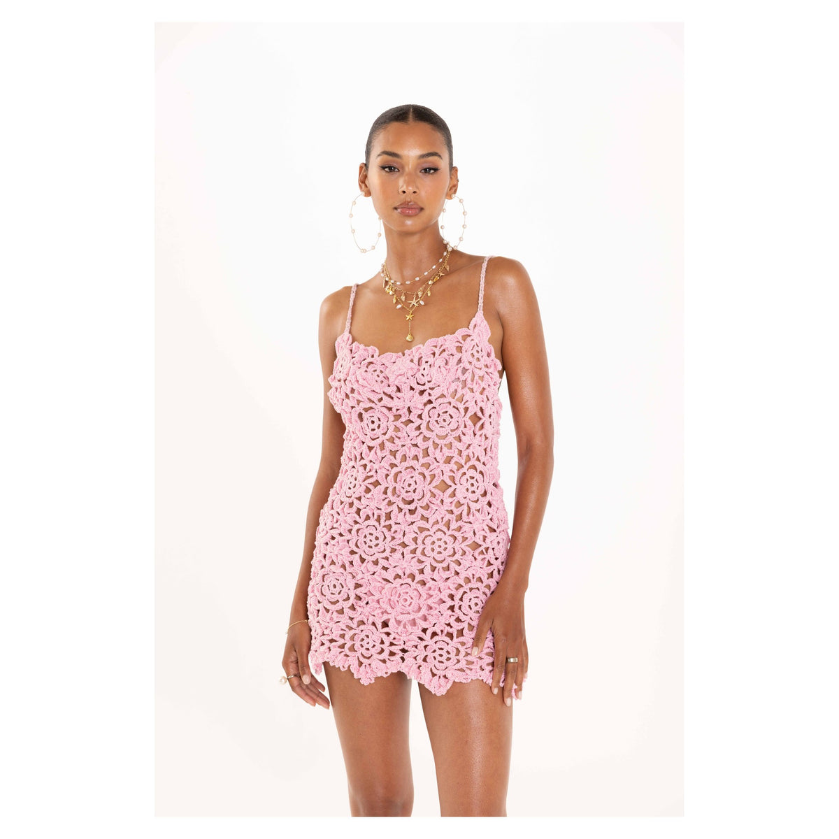 A woman stands against a white background wearing the ANDREEVA Pink Pearl Handmade Crochet Mini Dress—sleeveless, light pink, adorned with floral patterns in mercerized Brazilian cotton—styled with gold hoop earrings and layered necklaces.