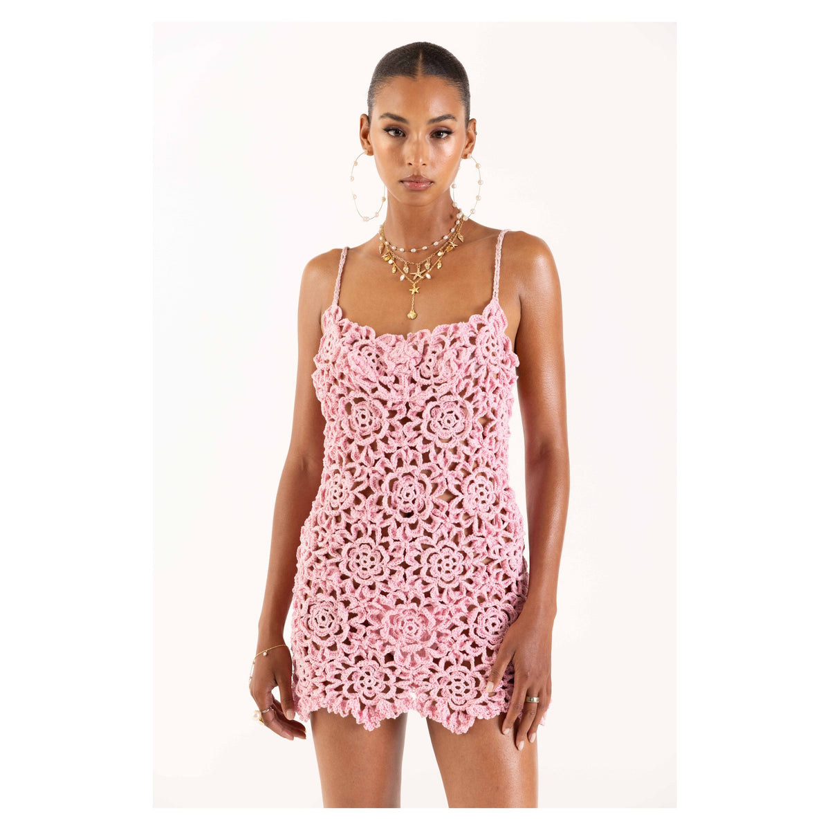A woman wears the ANDREEVA Pink Pearl Handmade Crochet Mini Dress, featuring a sleeveless design with floral patterns, against a plain white background. She accessorizes with hoop earrings, layered necklaces, and has her hair tied back.