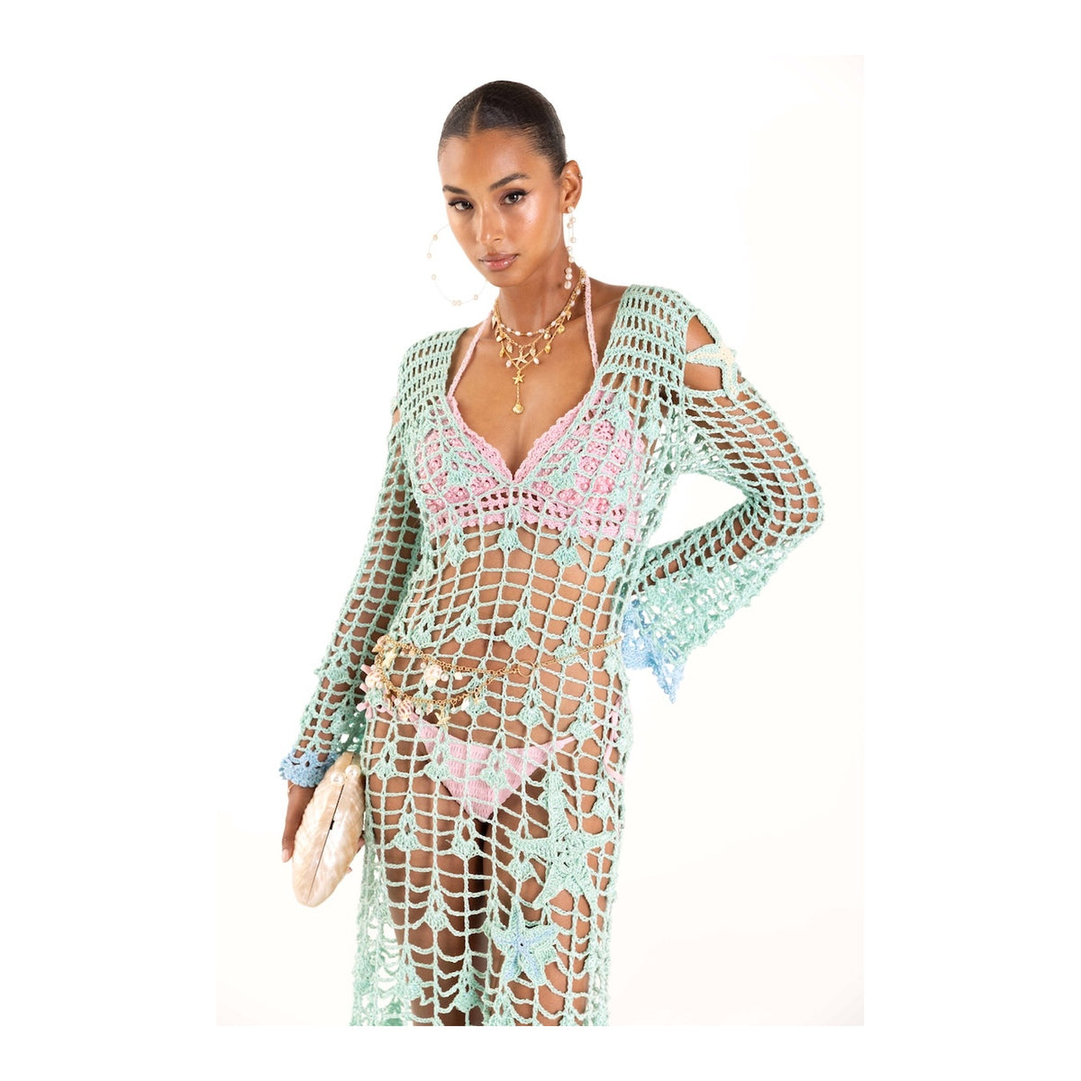 A woman poses confidently against a white background in the ANDREEVA Aqua Marine Handmade Maxi Cover-Up Dress over a pink bikini, accessorized with large hoop earrings, layered necklaces, and a small clutch.