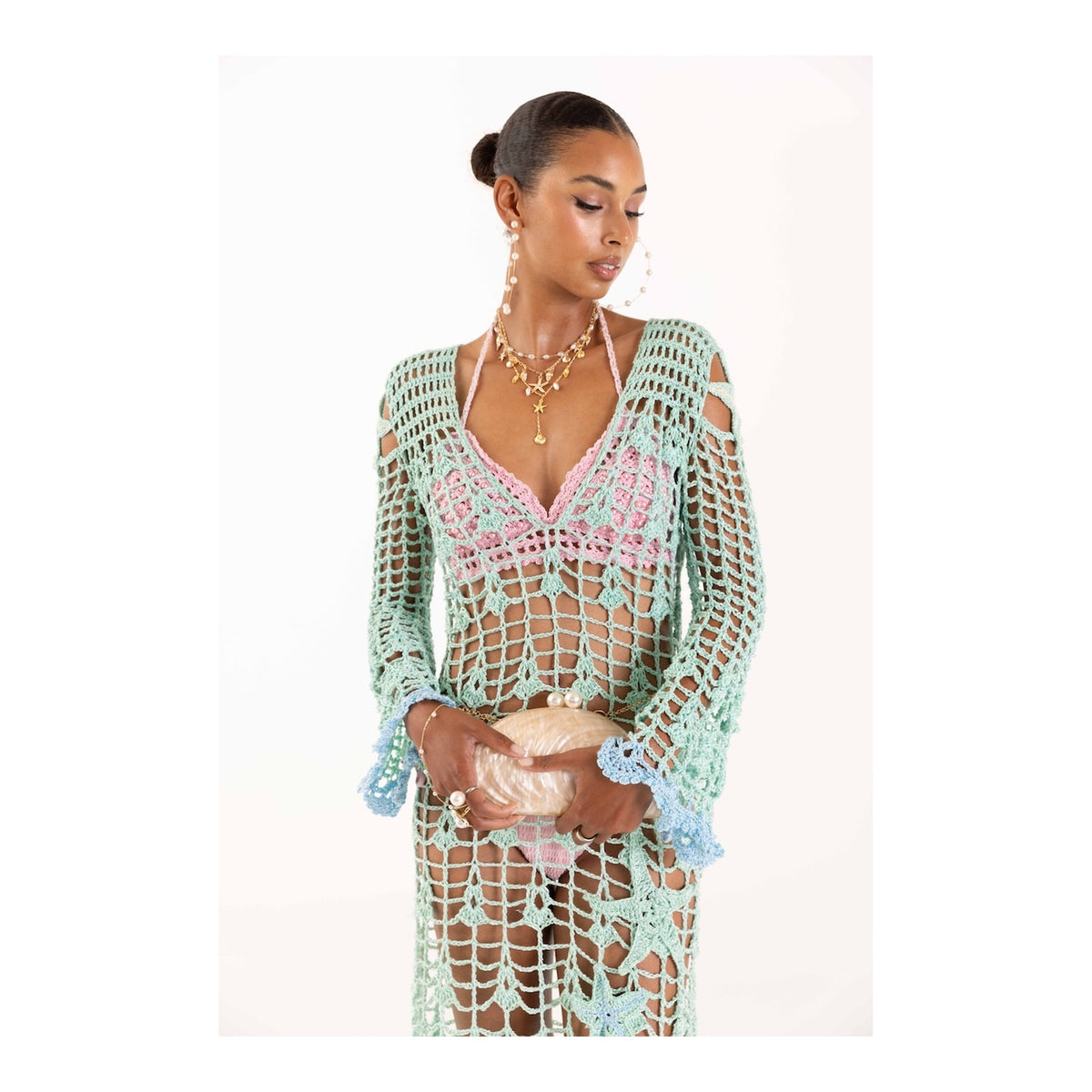 A woman wears the ANDREEVA Aqua Marine Handmade Maxi Cover-Up Dress in mint green over a pink bikini, holding a seashell-shaped bag. Her hair is in a sleek bun with layered gold necklaces, set against a white background.