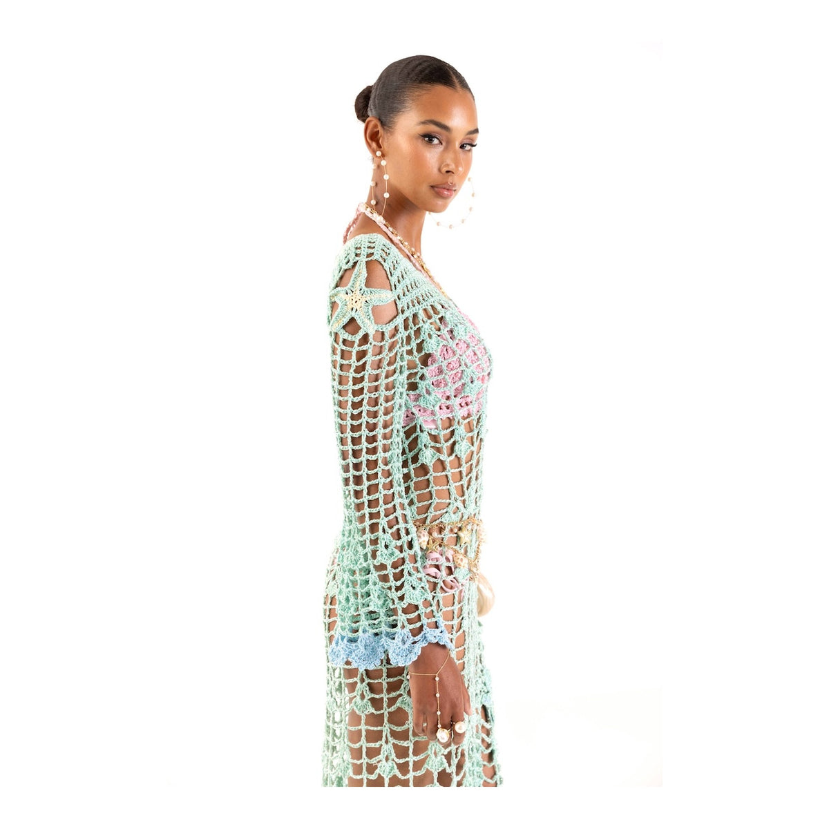 A woman stands in profile against a white background, wearing the ANDREEVA Aqua Marine Handmade Maxi Cover-Up Dress. Her hair is styled in a sleek bun, paired with large hoop earrings and a beaded belt.