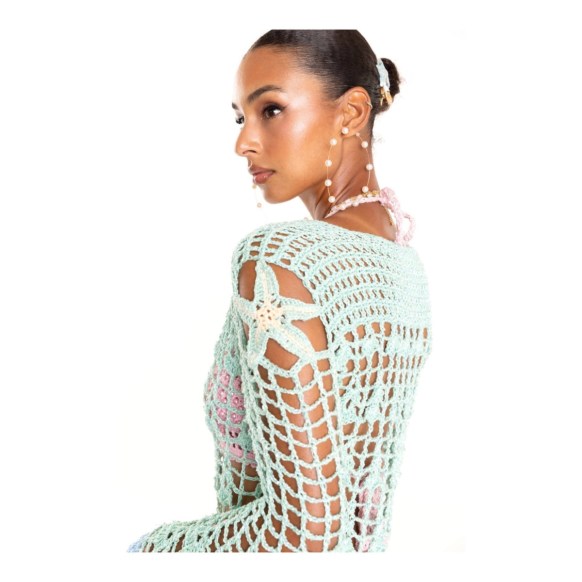 A woman with her hair in a bun models the ANDREEVA Aqua Marine Handmade Maxi Cover-Up Dress, featuring an open crochet pattern and floral details. She accessorizes with pearl hairpins and a beaded necklace, posing against a white background.