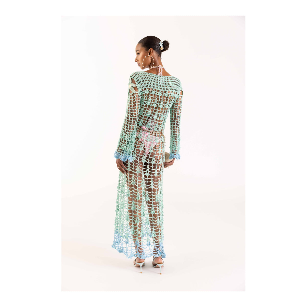 A woman faces away, wearing the ANDREEVA Aqua Marine Handmade Maxi Cover-Up Dress of Brazilian cotton with embroidery, styled over pastel layers, pearls in her hair and high heels, set against a plain white background.