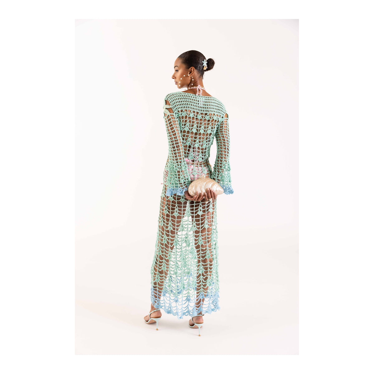 A woman stands facing away, wearing the ANDREEVA Aqua Marine Handmade Maxi Cover-Up Dress with a matching bikini. She holds a seashell-shaped clutch, dons high-heeled sandals, and her hair is styled in a neat bun against a white background.
