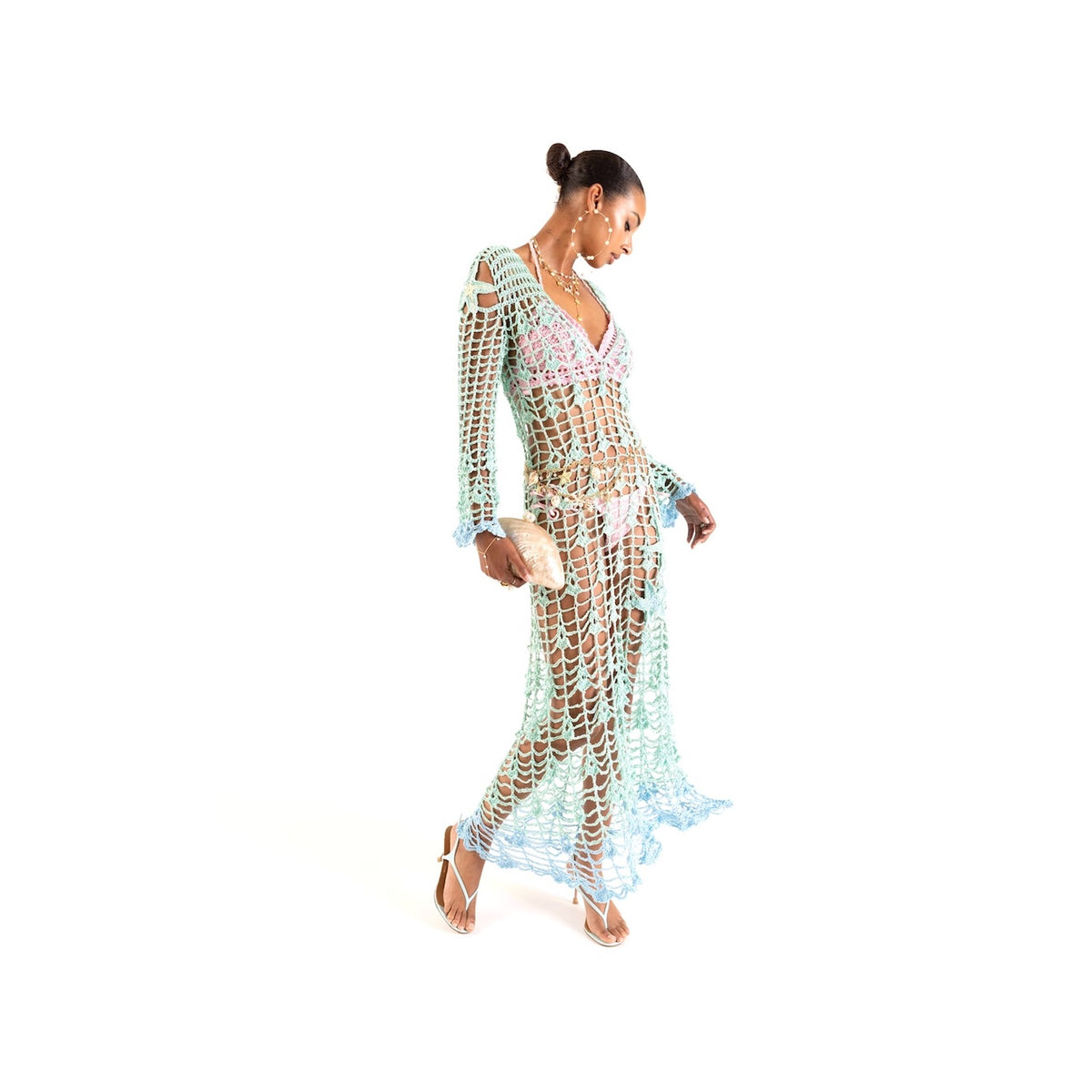 A woman in high-heeled sandals wears the ANDREEVA Aqua Marine Handmade Maxi Cover-Up Dress of pastel mesh crochet over a bikini, with handmade embroidery. She holds a small beige clutch, looking down with one knee bent against a white background.