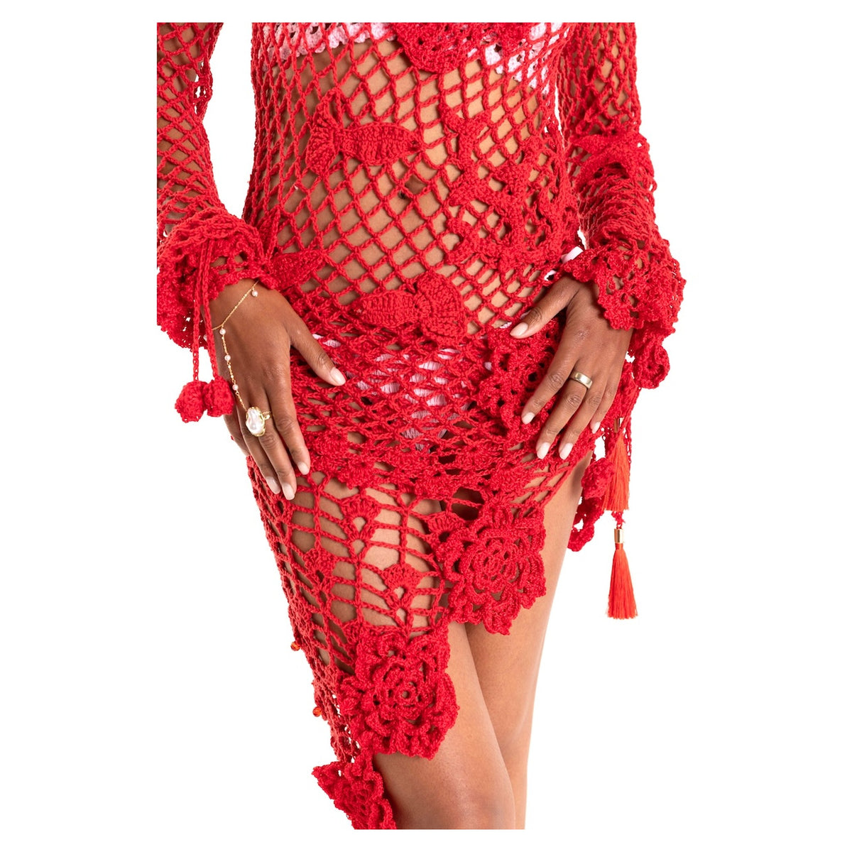 A person models the ANDREEVA Cherry Handmade Crochet Cover-Up, featuring red floral patterns and tassel details. The artisanal dress reveals the midsection, while their hands are accessorized with rings and bracelets.
