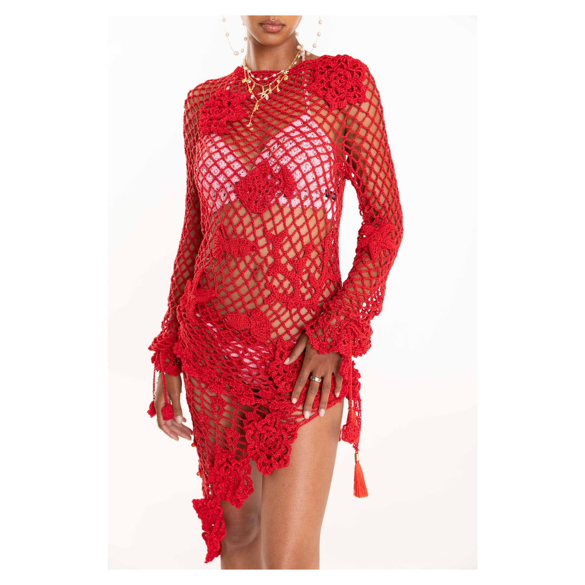 A woman wears the ANDREEVA Cherry Handmade Crochet Top, a sheer red cotton cover-up with floral crochet, long sleeves, asymmetrical hem, tassels, and textured details over a red bikini—ideal for beach layering.