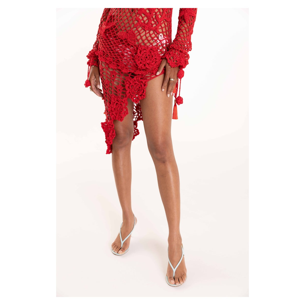 A model showcases the ANDREEVA Cherry Handmade Crochet Cover-Up, a red floral-patterned dress with long sleeves, on a white background. The look is styled with white heeled sandals; the model's face is not visible.