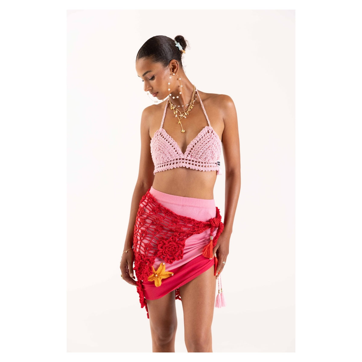A woman stands against a plain background wearing the ANDREEVA Pink Pearl Handmade Crochet Bra, paired with a pink mini skirt and red fishnet wrap with a yellow flower accent. She finishes her chic resort look with layered necklaces, earrings, and a low bun.
