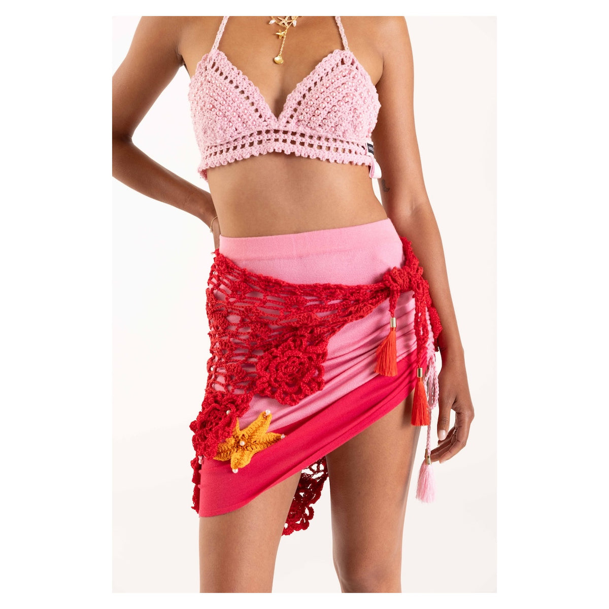 A woman models the Pink Pearl Handmade Crochet Bra by ANDREEVA, paired with a matching pink skirt featuring a red crochet overlay, floral accents, and a yellow starfish ornament—ideal for chic resort wear. Only her torso and upper legs are visible.