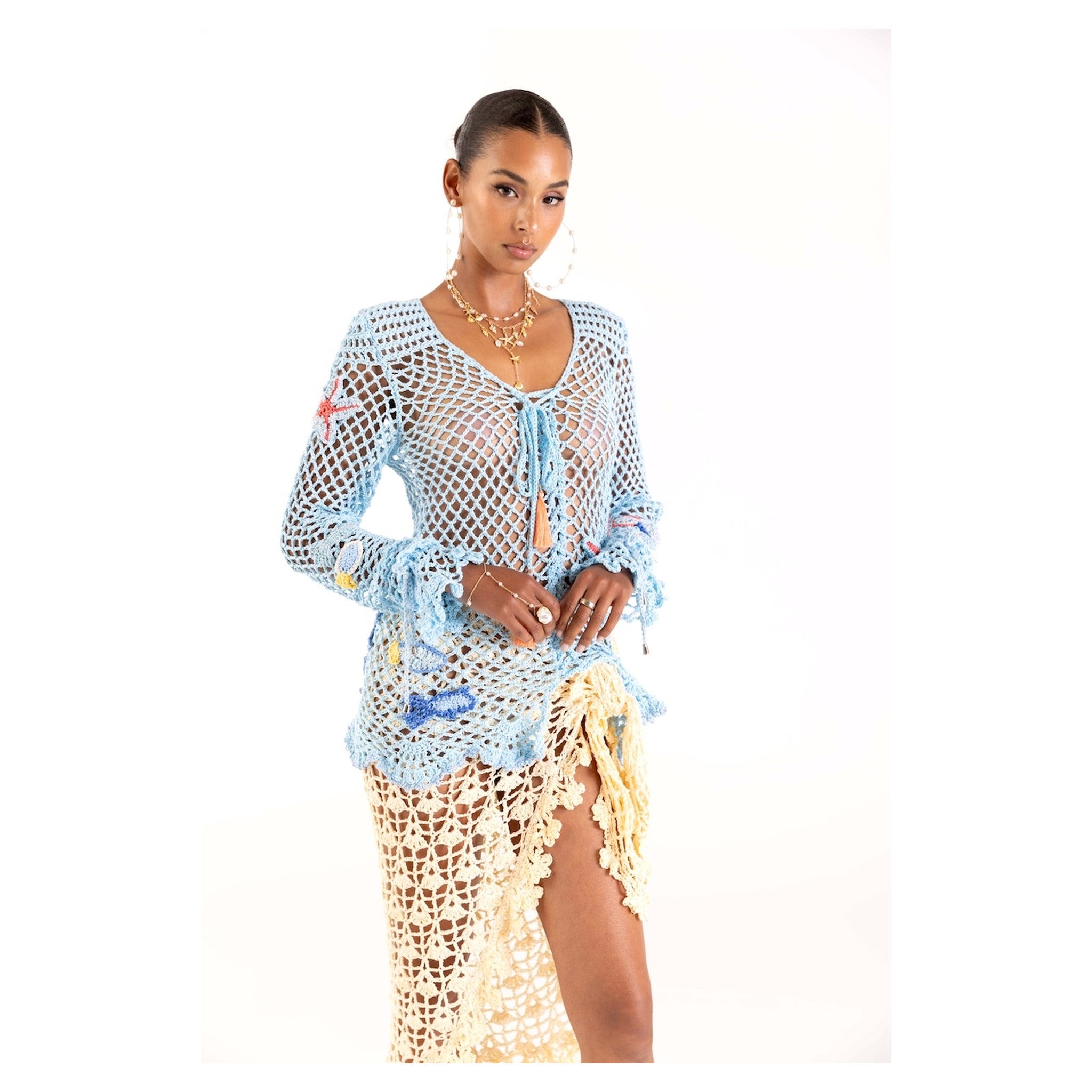 A woman poses against a white background in the ANDREEVA Aqua Marine Handmade Crochet Top-Cover Up, layered over a yellowish cream crochet skirt with a high slit. She completes the look with layered gold necklaces and earrings.