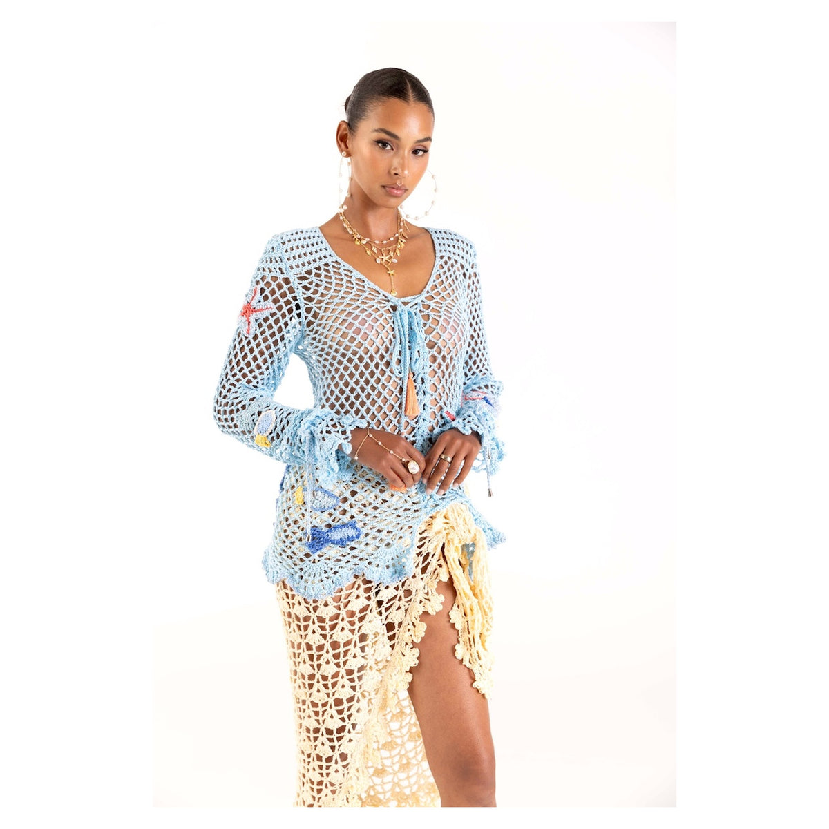 A woman poses against a white background in the ANDREEVA Aqua Marine Handmade Crochet Top-Cover Up, layered over a yellowish cream crochet skirt with a high slit. She completes the look with layered gold necklaces and earrings.