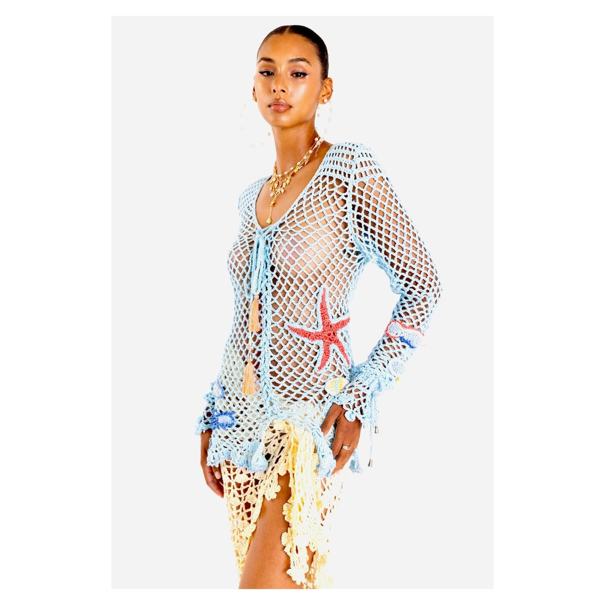 A woman models the ANDREEVA Aqua Marine Handmade Crochet Top-Cover Up, featuring a sheer, light blue fishnet design with colorful sea-themed details over a nude bikini, accessorized with gold jewelry against a white background.