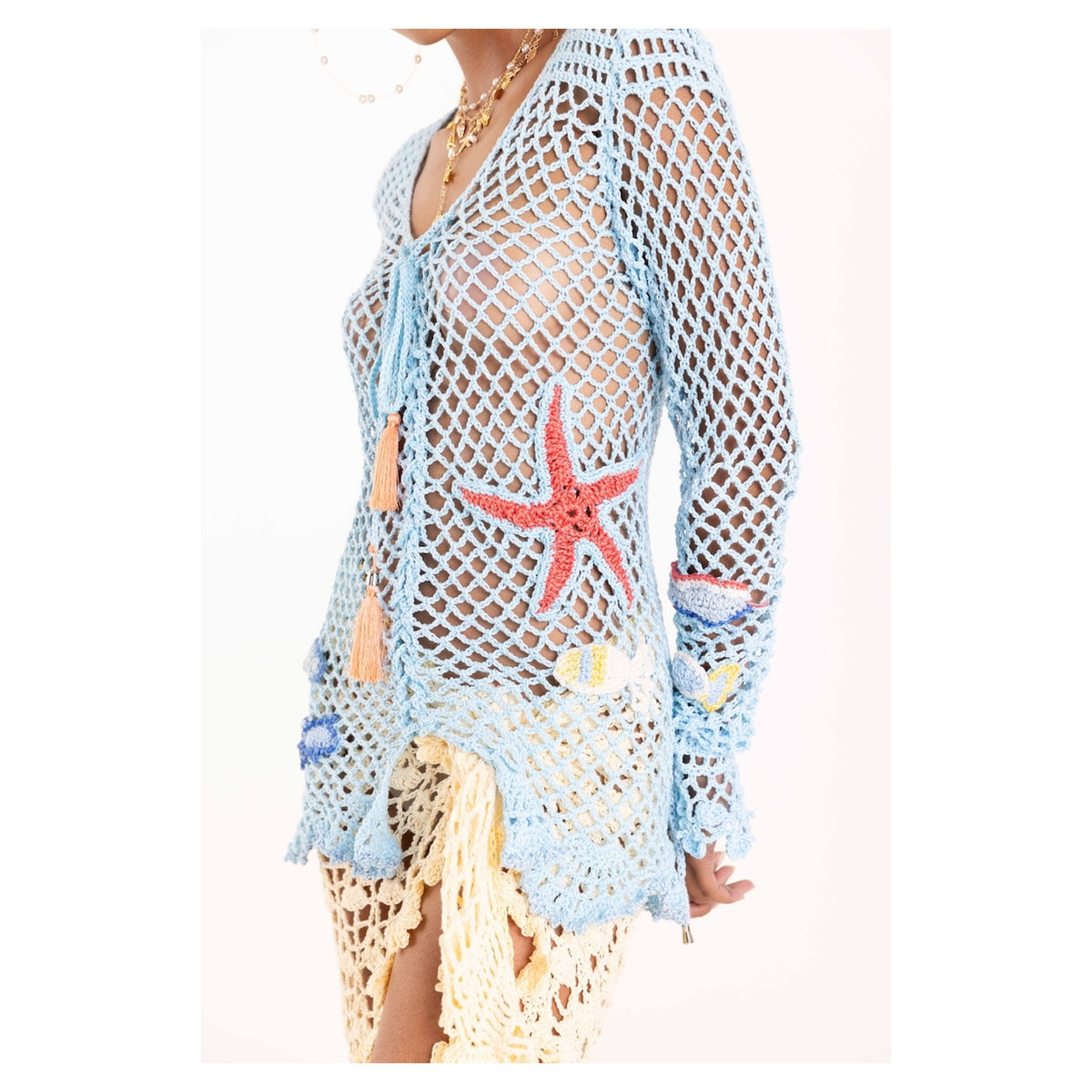 A person wears the ANDREEVA Aqua Marine Handmade Crochet Top-Cover Up with colorful sea-themed appliqués, layered over a pale yellow skirt—perfect for a playful beach look.