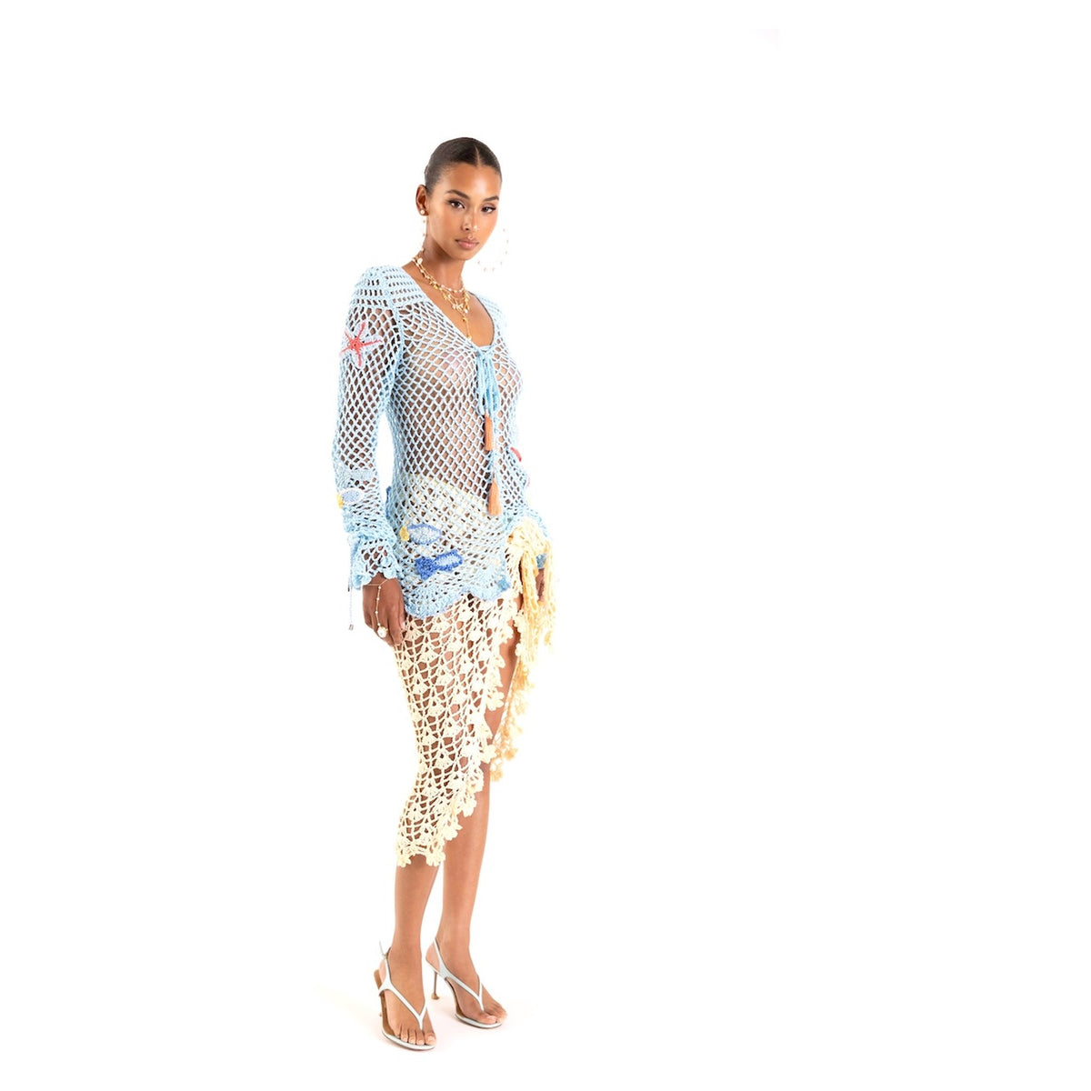 A woman models the ANDREEVA Beach Rose Handmade Crochet Cover-Up, featuring blue and cream patterns, paired with white strappy heels against a plain white background. She gazes at the camera with a neutral expression.