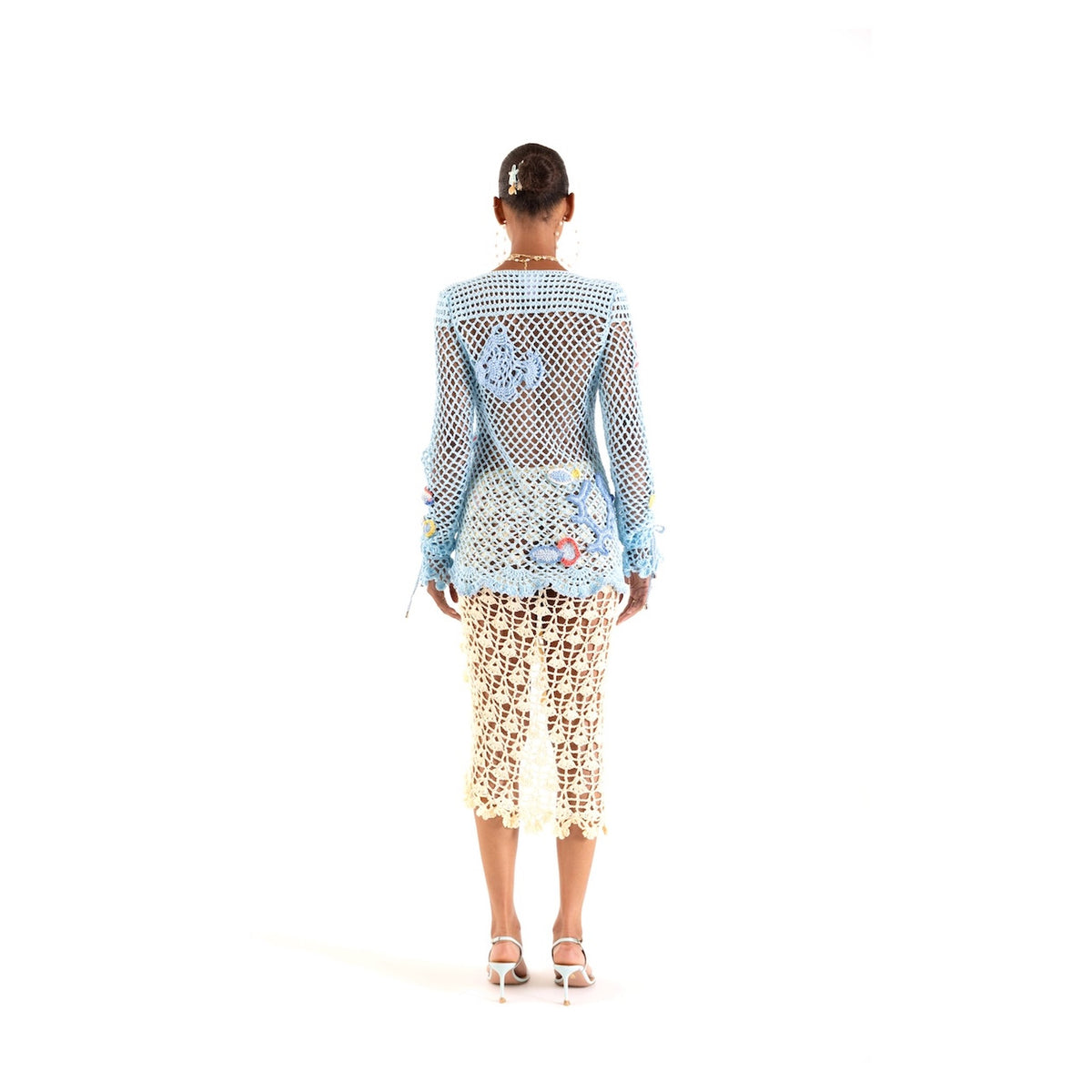 A woman faces away, wearing the ANDREEVA Aqua Marine Handmade Crochet Top-Cover Up with a sheer blue mesh layer featuring butterfly designs. Her hair is in a bun with a flower clip, and she pairs white high-heeled sandals against a plain white background.