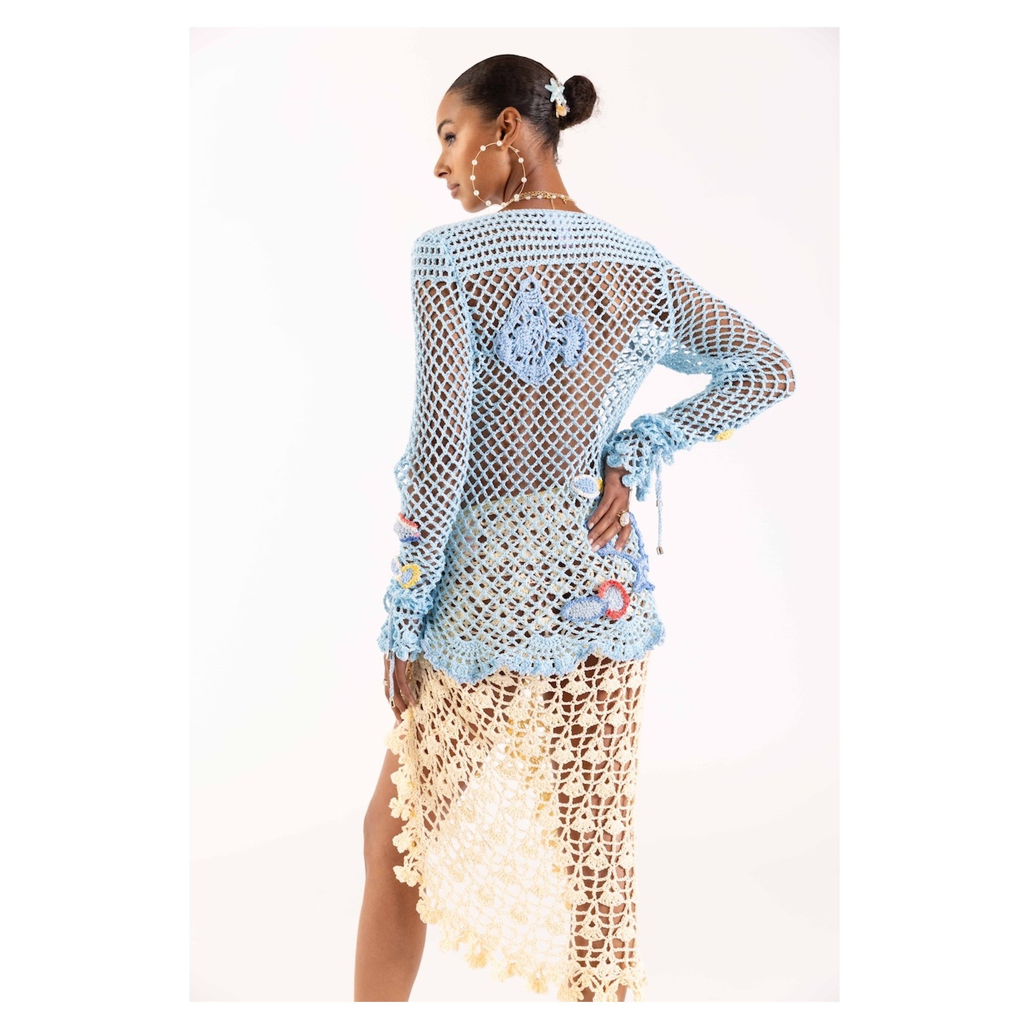 A woman faces away from the camera, wearing the ANDREEVA Aqua Marine Handmade Crochet Top-Cover Up in sheer light blue cotton with floral patterns, paired with a cream tassel skirt. She completes her look with hoop earrings and a hair accessory.