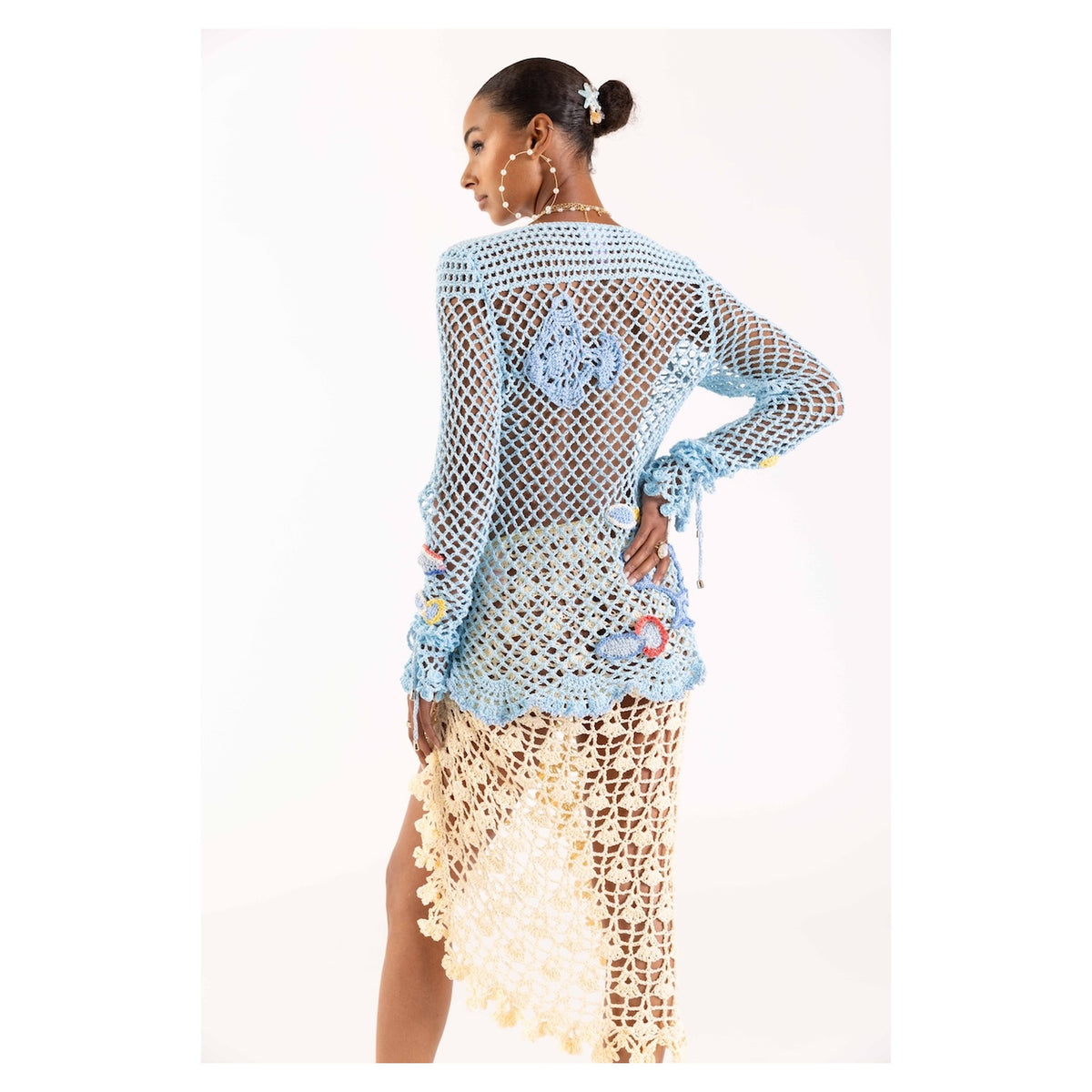A woman faces away from the camera, wearing the ANDREEVA Aqua Marine Handmade Crochet Top-Cover Up in sheer light blue cotton with floral patterns, paired with a cream tassel skirt. She completes her look with hoop earrings and a hair accessory.