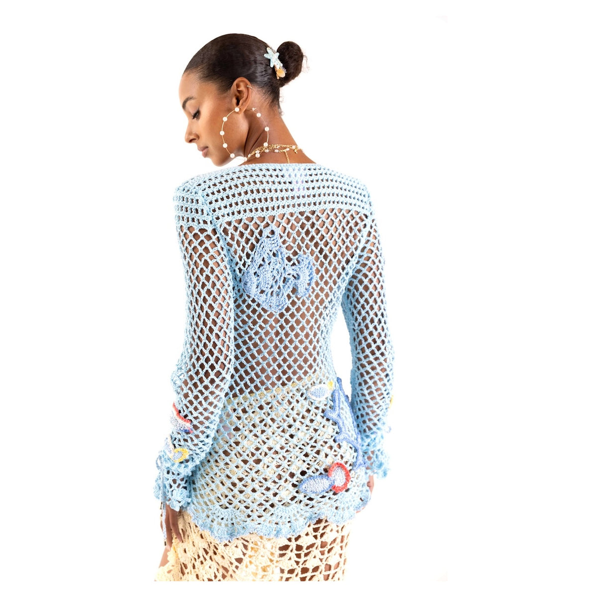 A woman with a slicked-back bun and hair clip models the ANDREEVA Aqua Marine Handmade Crochet Top-Cover Up, light blue with floral details, seen from the back. She pairs it with gold hoop earrings and layered necklaces—ideal as a beach cover up.