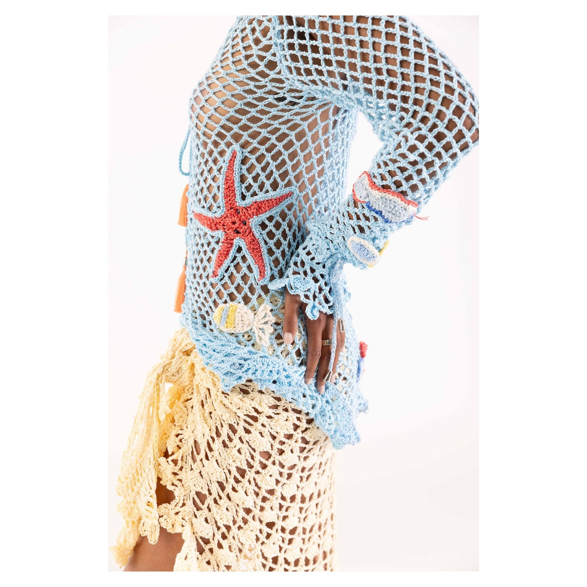 A person models the ANDREEVA Beach Rose Handmade Crochet Cover-Up, a blue fishnet top with red starfish and seashell appliqués, paired with a beige skirt. This unique beach-inspired resort wear makes a statement; only the body and hand are visible.