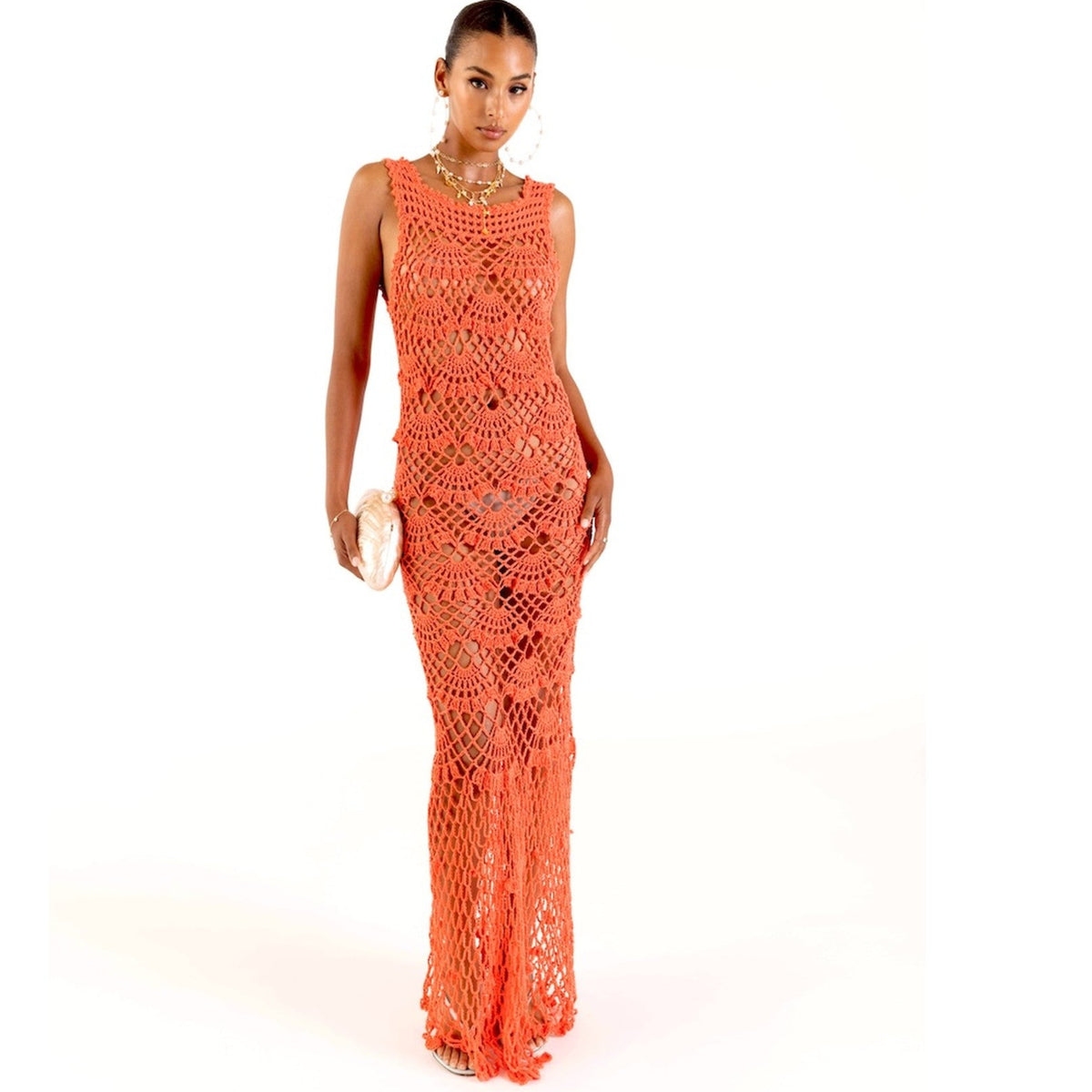 A woman models the ANDREEVA Coral Shell Handmade Maxi Crochet Dress, sleeveless and floor-length, paired with a clutch, gold hoop earrings, and a necklace against a white background for a coastal-chic look.