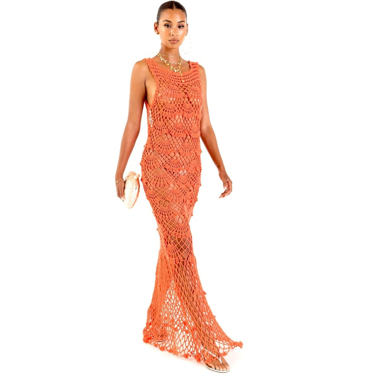 A woman wears the ANDREEVA Coral Shell Handmade Maxi Crochet Dress, styled with a beige clutch and gold jewelry, radiating coastal elegance against a white background.
