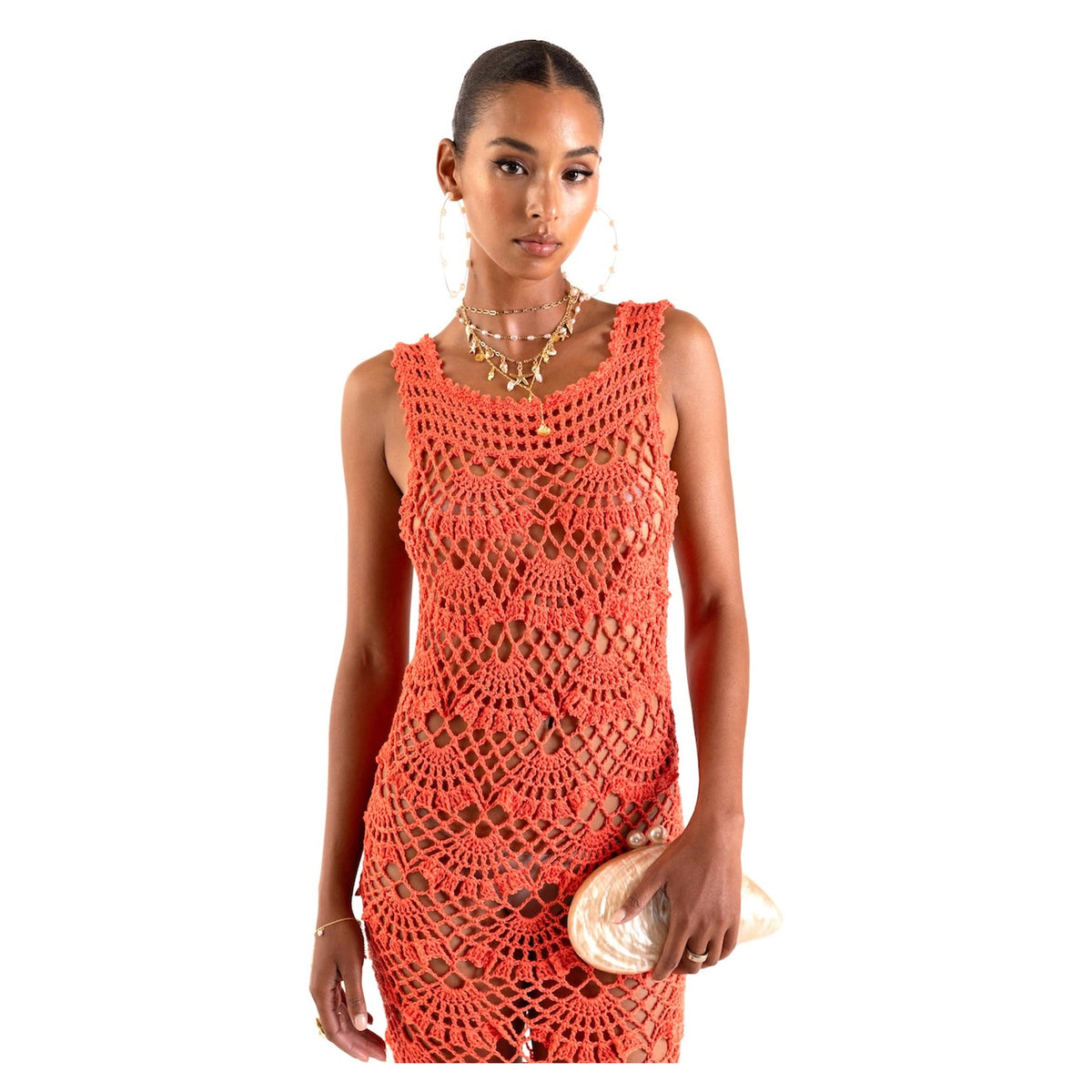 A woman models the ANDREEVA Coral Shell Handmade Maxi Crochet Dress, styled with a shiny light clutch, large hoop earrings, and layered necklaces for a touch of coastal elegance against a white background.