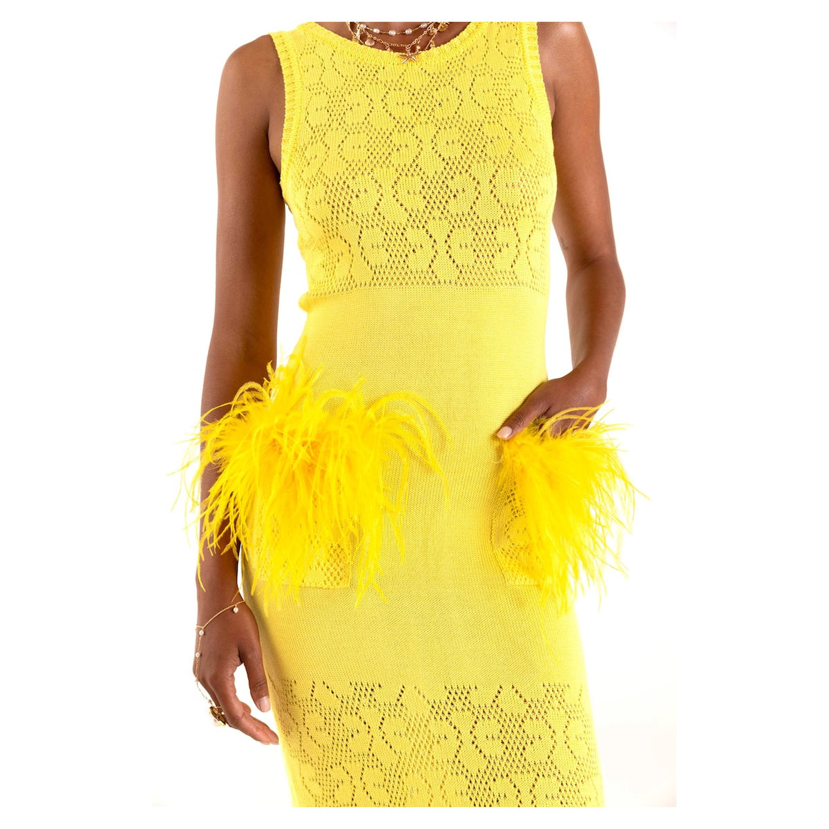 A person wearing the ANDREEVA Lemon Knit Dress, a sleeveless yellow knit dress featuring lace-like patterns and feather-accented hip pockets, styled with layered necklaces and bracelets.