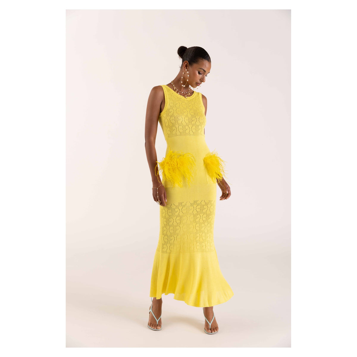 A woman stands against a plain light bamboo background wearing the ANDREEVA Lemon Knit Dress—sleeveless, floor-length with lace and feather accents—paired with strappy heels and a pearl necklace.