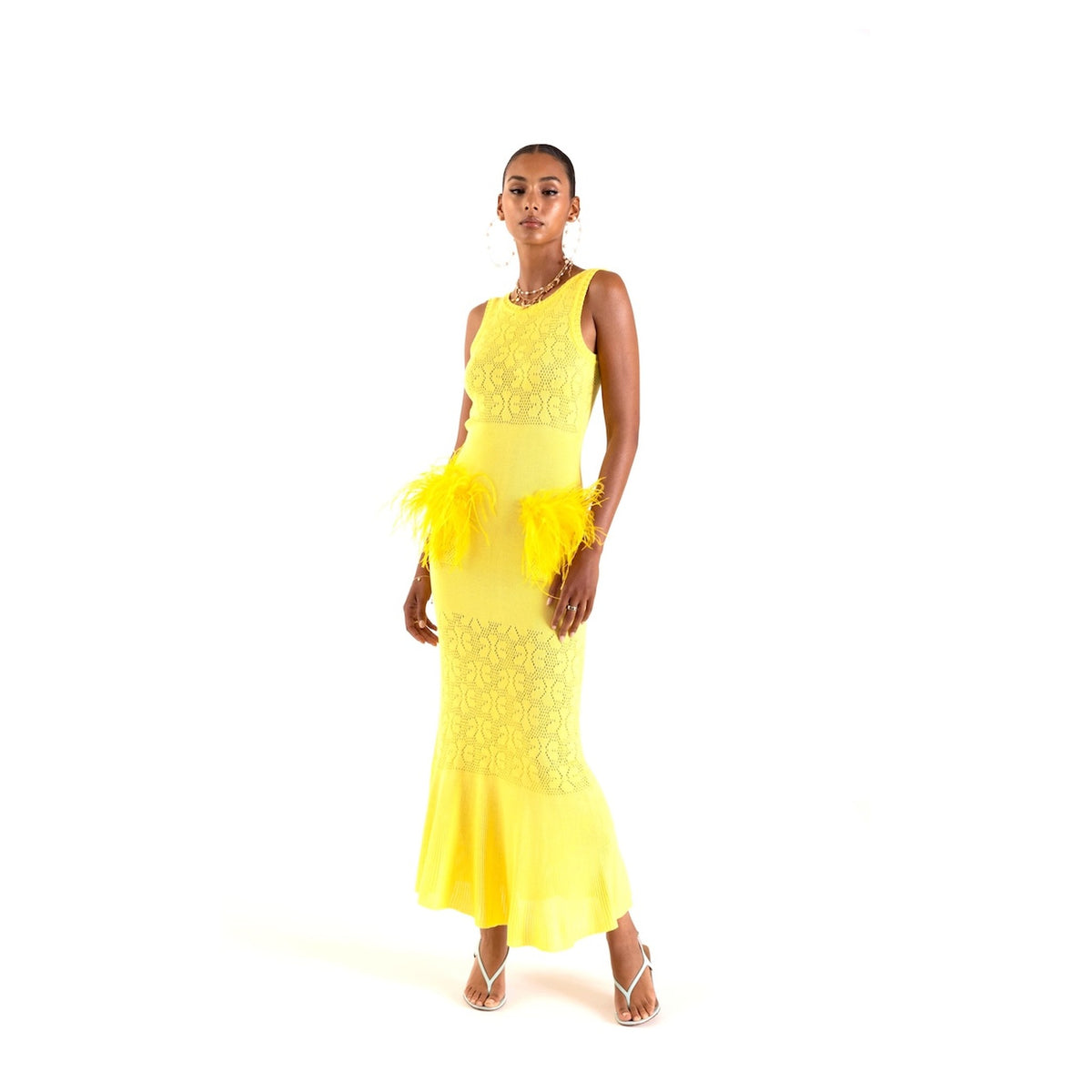 A woman models the ANDREEVA Lemon Knit Dress—an ankle-length, sleeveless yellow knit dress with feather accents at the hips—paired with gold earrings, a necklace, and heeled sandals, set against a plain white background.