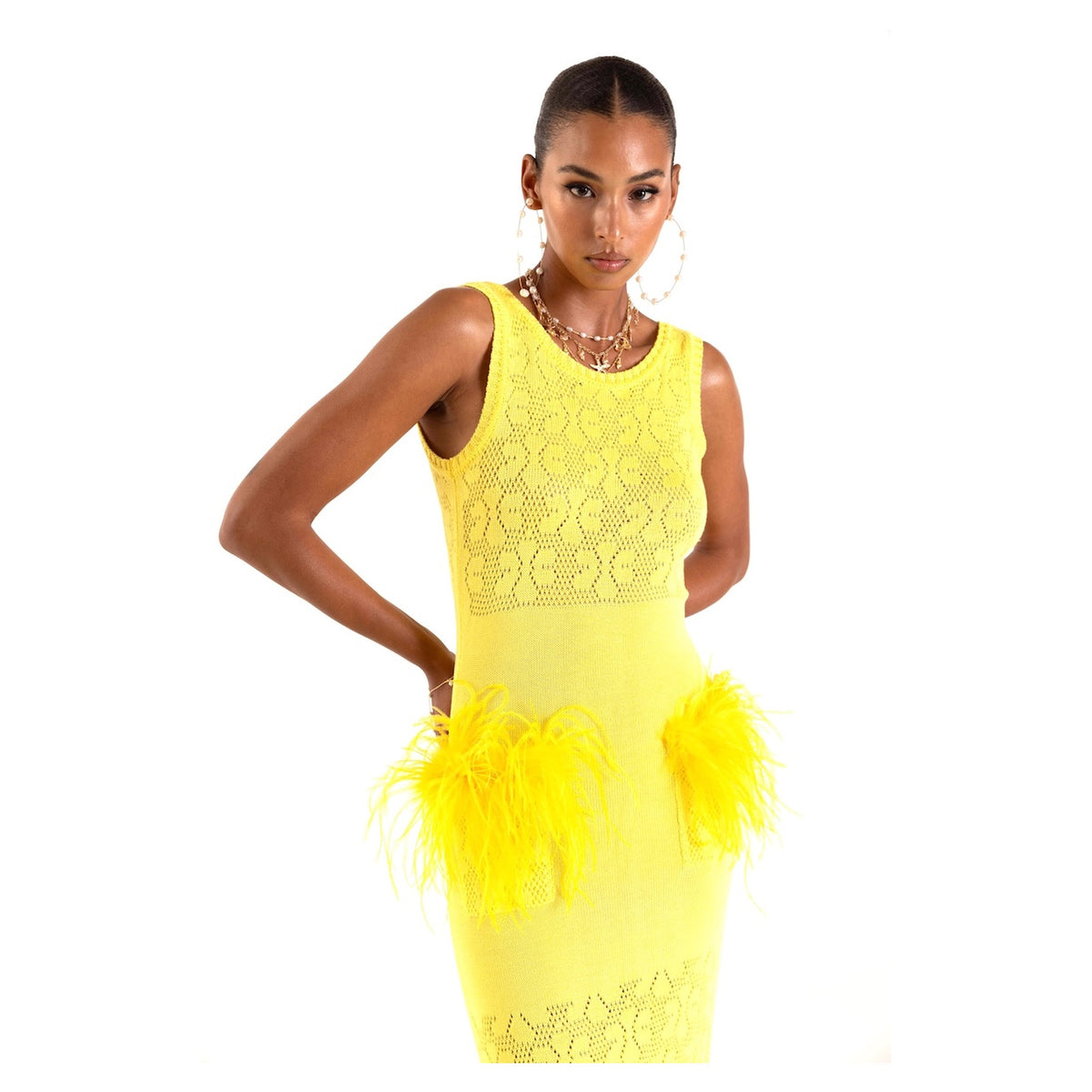 A woman poses beside slender bamboo stalks against a white background, wearing the ANDREEVA Lemon Knit Dress—sleeveless, bright yellow with lace and feather hip accents—styled with large hoop earrings and layered necklaces.