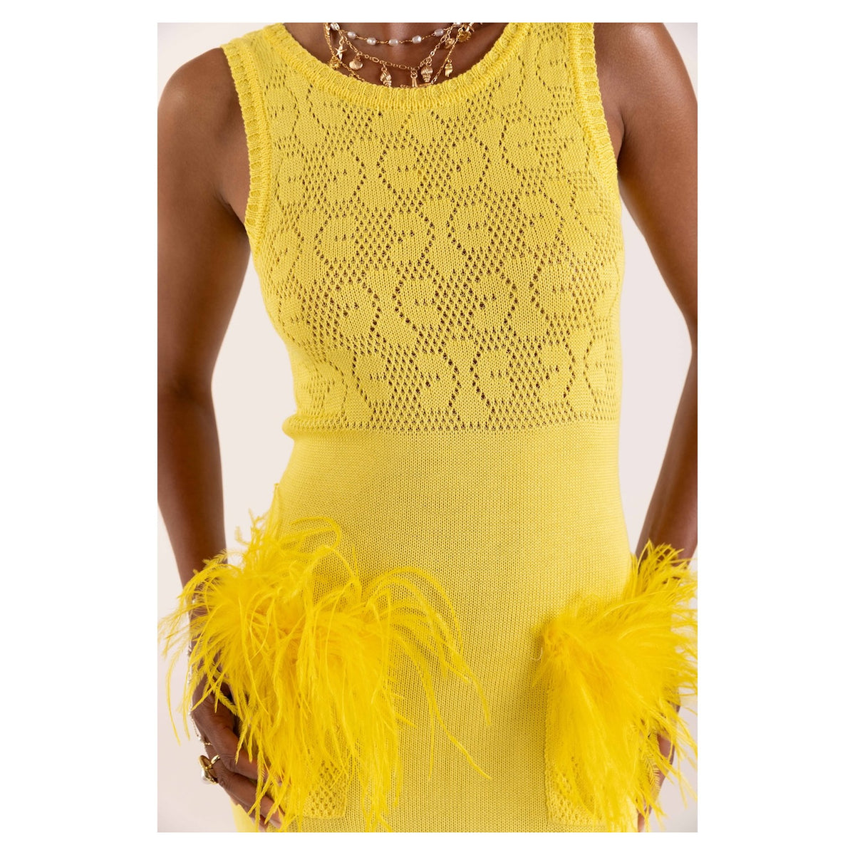 A person wears the ANDREEVA Lemon Knit Dress, a sleeveless bright yellow knit with a textured pattern and bold feather accents at the hips. Gold necklaces are layered around the neck. The photo is cropped from shoulders to knees.