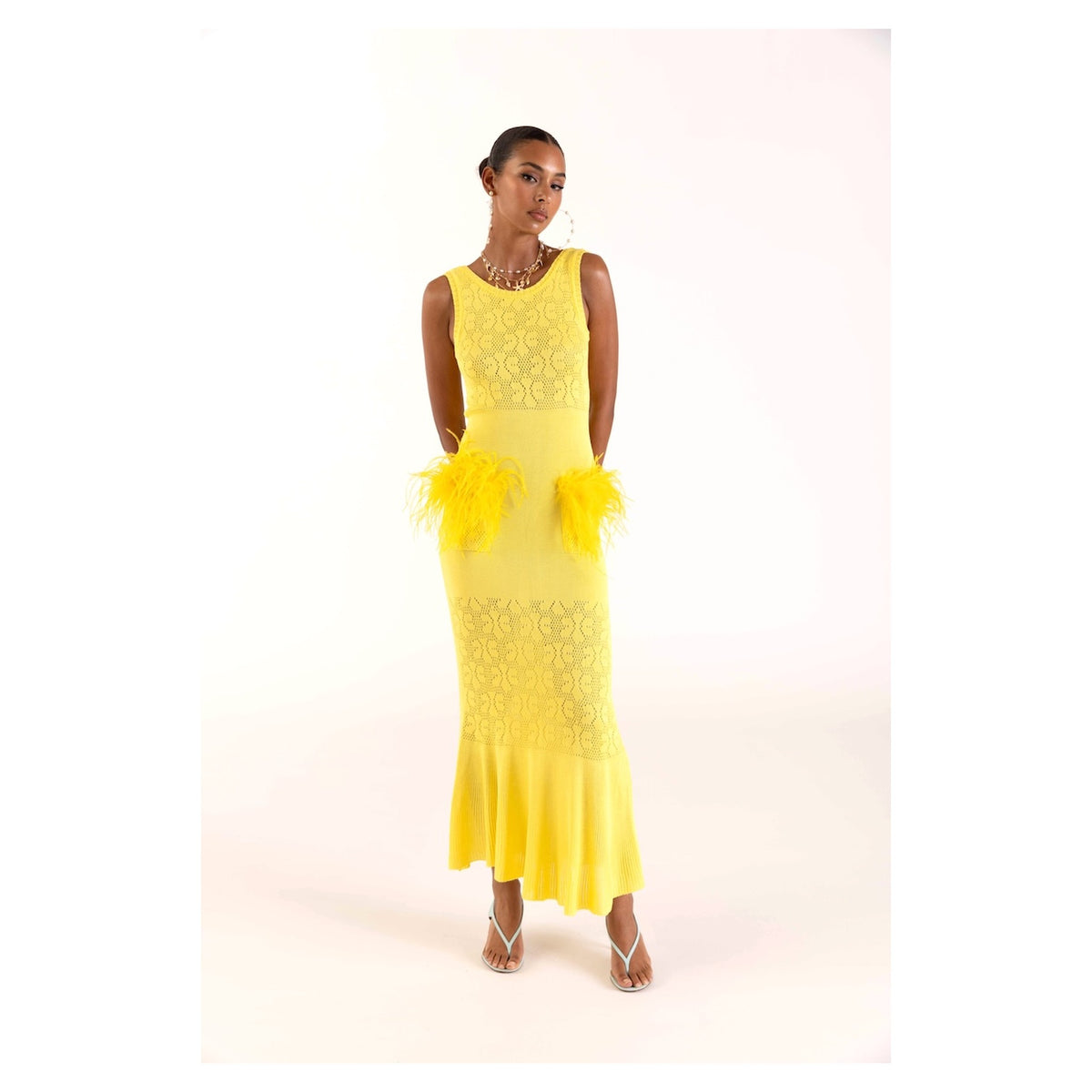 A woman poses against a white background in the ANDREEVA Lemon Knit Dress, a sleeveless, ankle-length yellow dress with feather accents at the hips, styled with strappy heels, earrings, and a necklace.