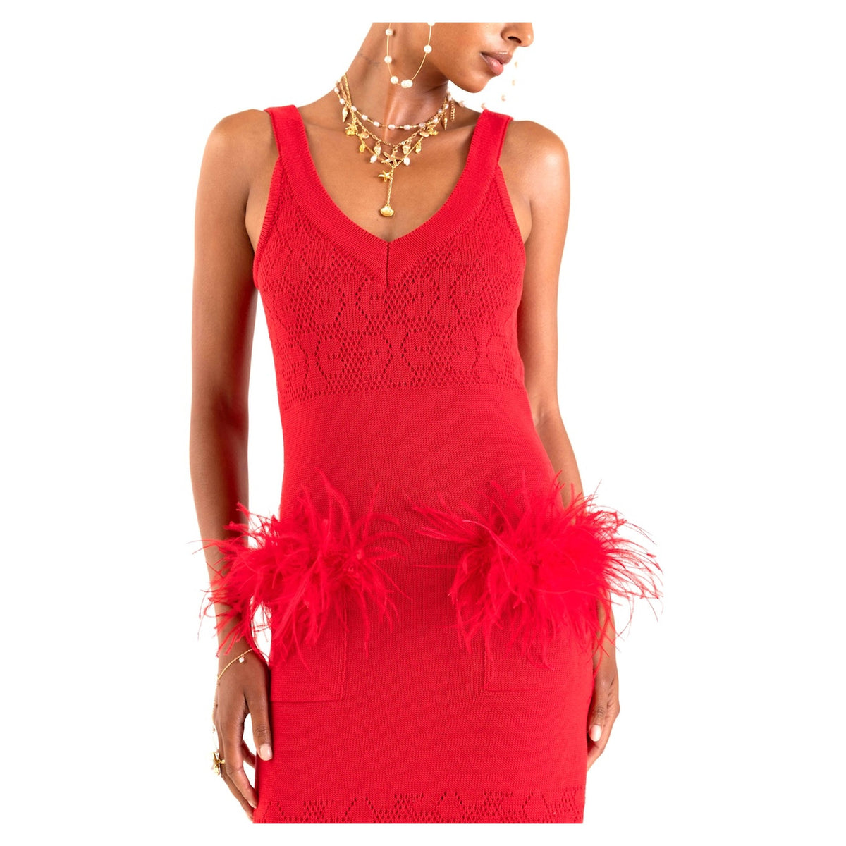 A woman wears the ANDREEVA Cherry Knit Dress With Feather Details—a sleeveless, V-neck textured knit dress featuring two large feather accents at the hips, accessorized with layered gold necklaces and earrings.