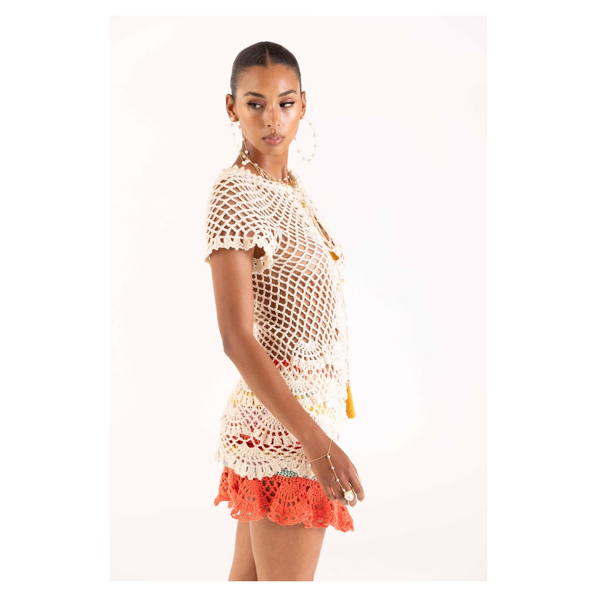 A woman poses in the ANDREEVA Coral Reef Handmade Crochet Mini Skirt, paired with a white crochet top and hoop earrings, looking to her left against a plain white background.