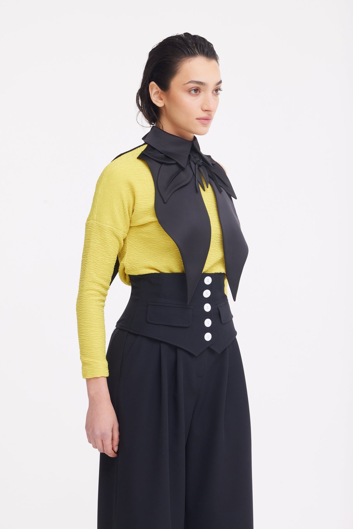 A woman stands against a plain background wearing a yellow textured long-sleeve top, a large black bow around her shoulders and neck, and JULIA ALLERT’s Buttoned Corset-Waist Black Tailored Trousers.