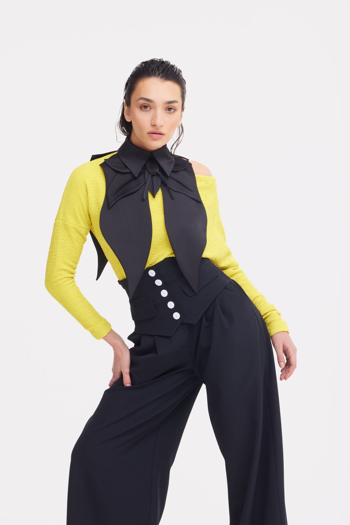 A woman poses confidently in a bright yellow off-shoulder top, styled with JULIA ALLERT’s Buttoned Corset-Waist Black Tailored Trousers and a black scarf tied in a bow. The background is plain white.