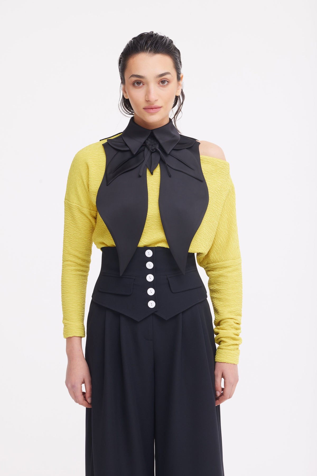 A woman stands against a white background wearing JULIA ALLERT's Buttoned Corset-Waist Black Tailored Trousers, a yellow off-shoulder top, and a large black bow tied around her neck.