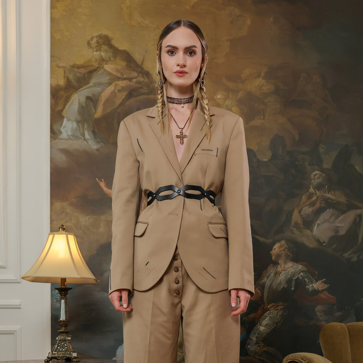 Woman in a beige suit standing in a room with classical painting and lamp.