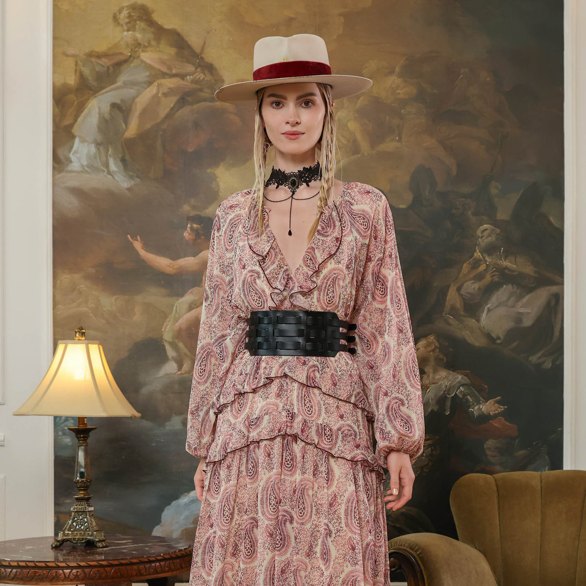 Woman in a patterned dress and hat standing in a room with a large painting in the background.