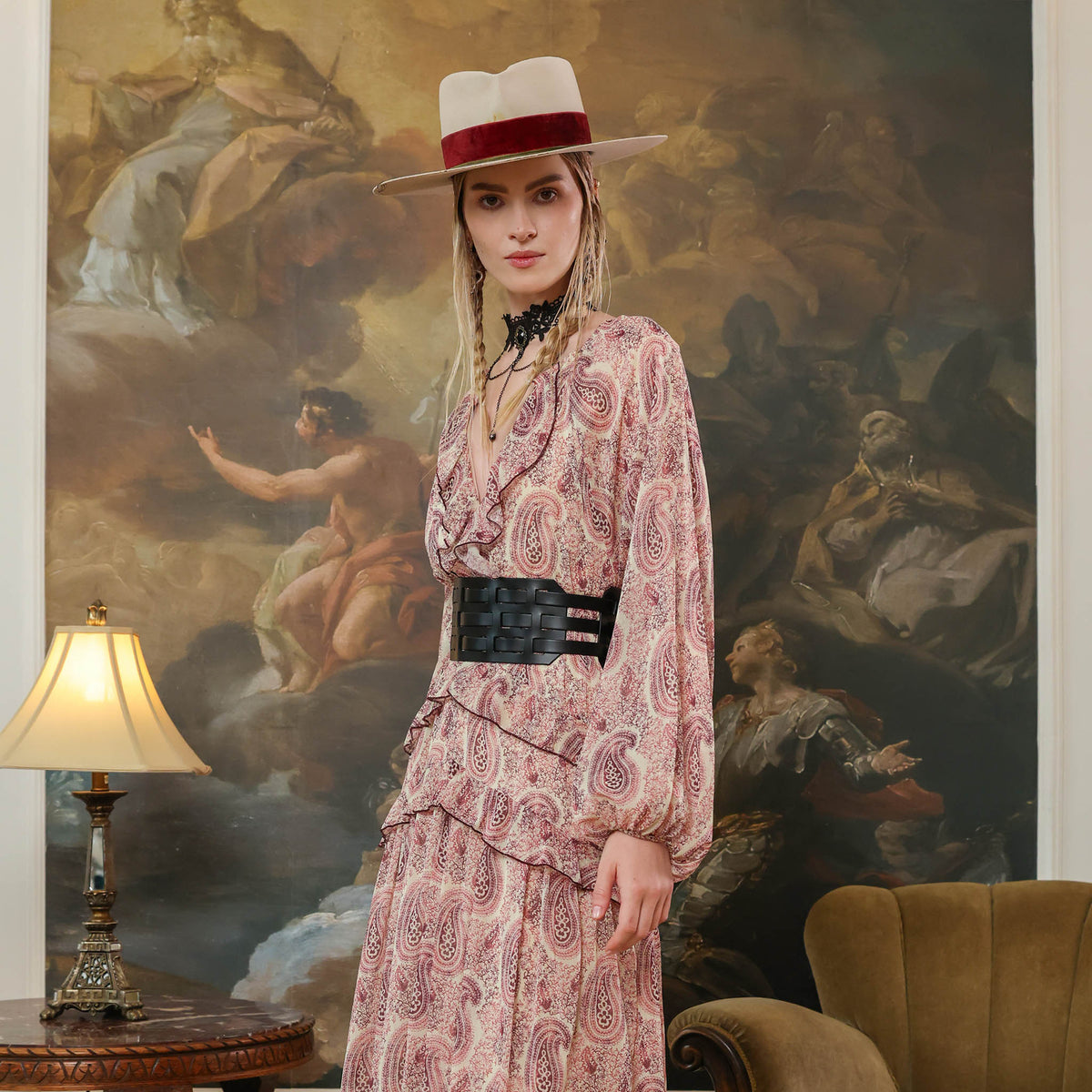 Woman in a patterned dress and hat standing in front of a classical painting