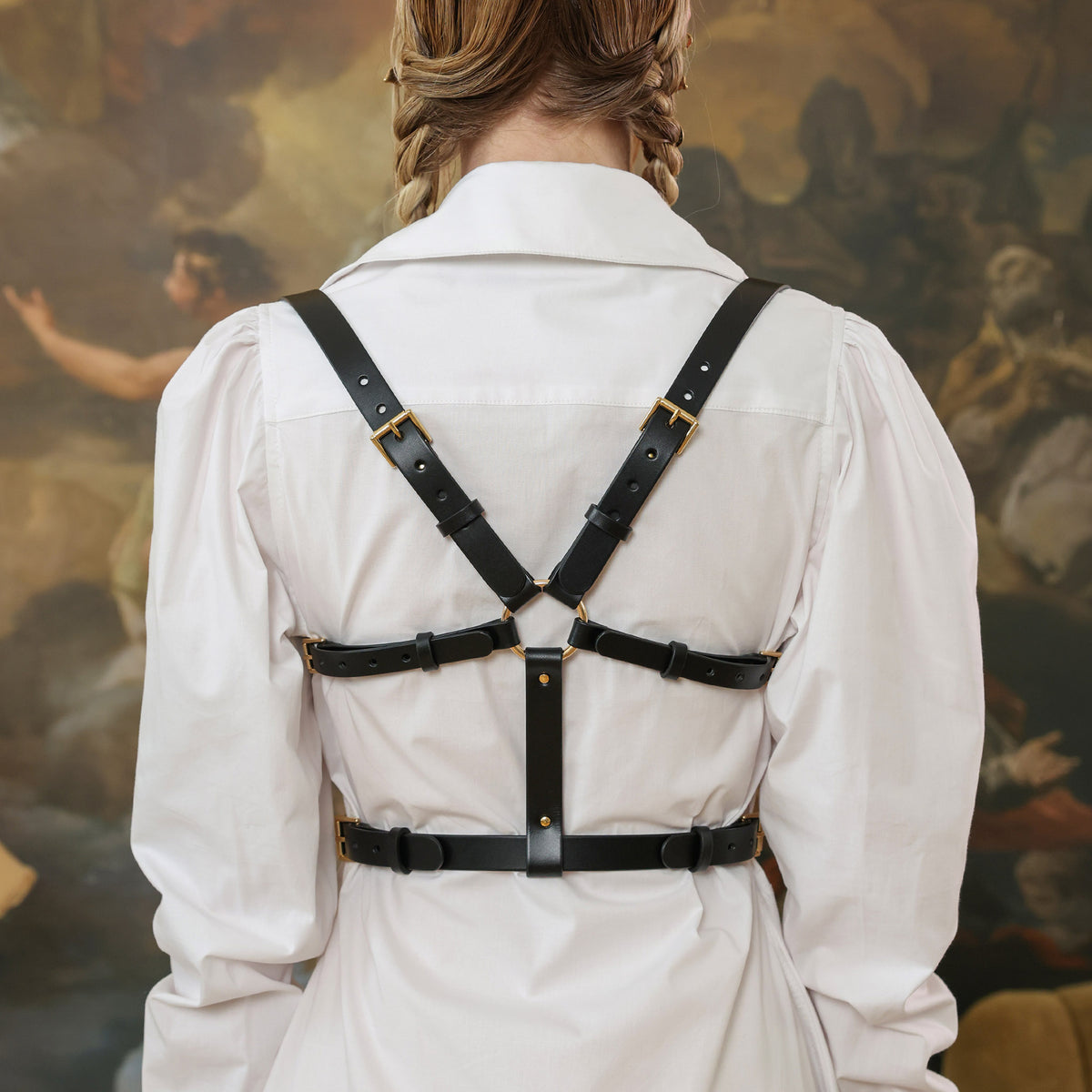 Person wearing a black leather harness over a white shirt with a blurred painting background