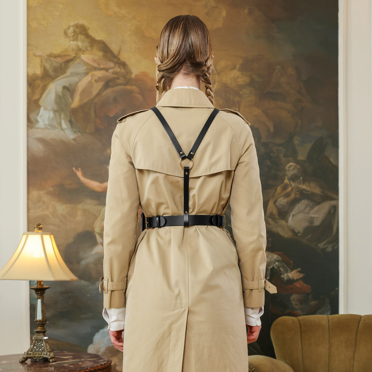 Woman in a beige trench coat standing in front of a classical, black leather harness with a belt and pouchpainting
